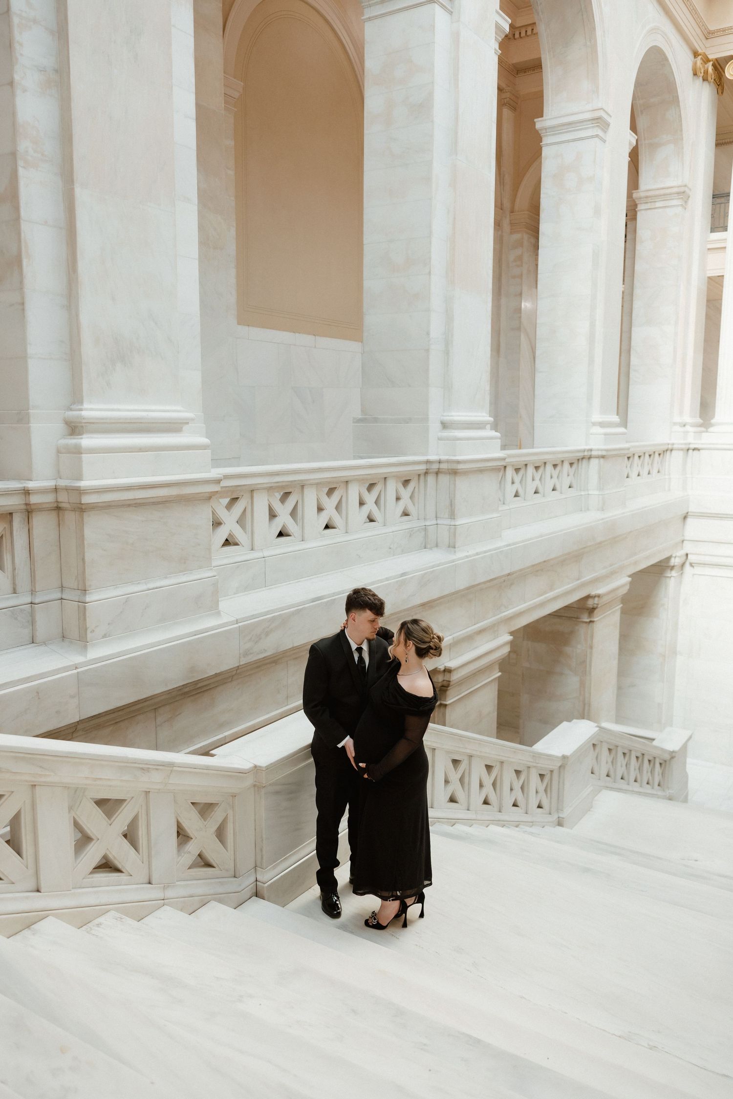 Couple in formal black attire sharing romantic moment on elegant white marble staircase interior.