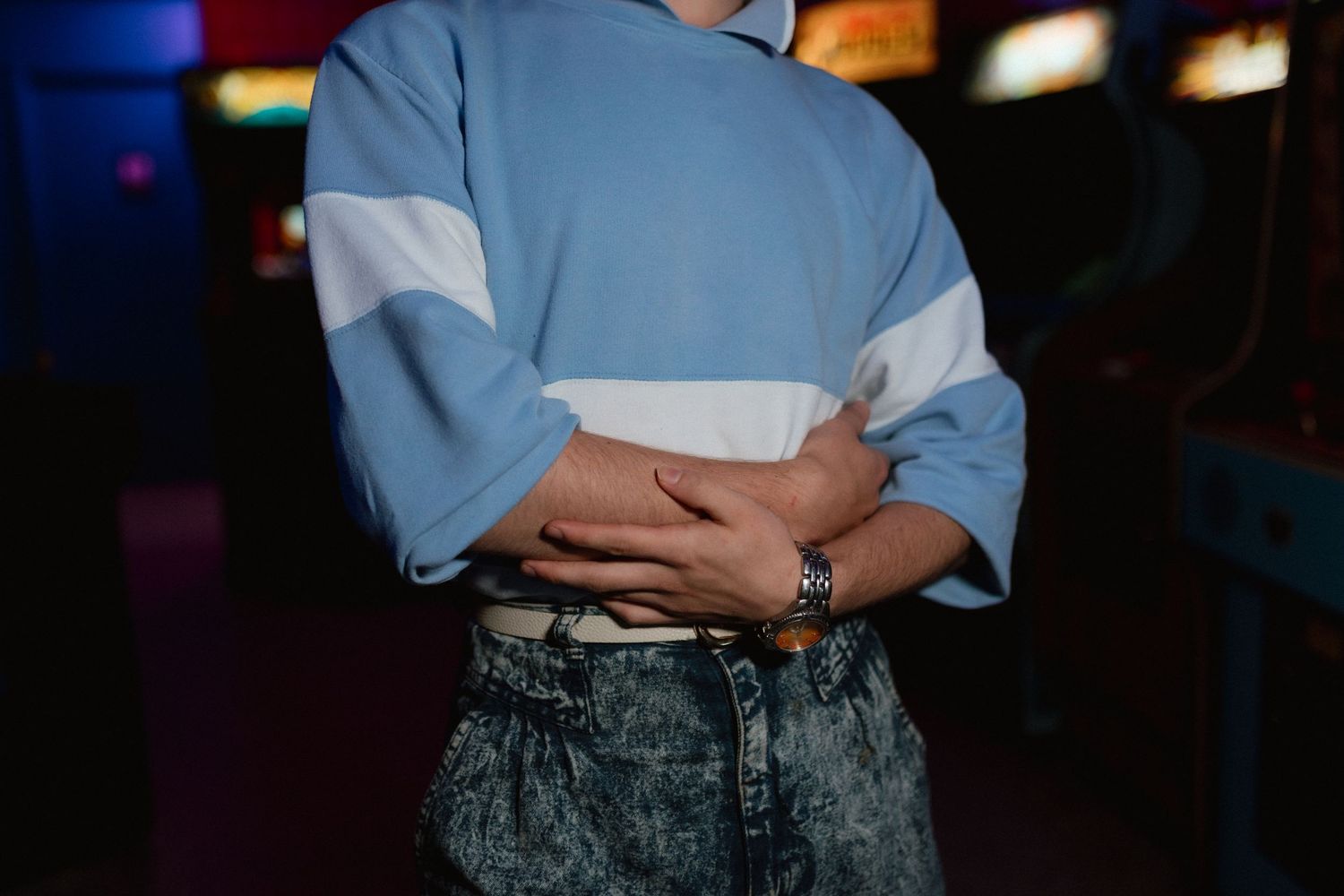 Close-up of a person in a blue and white retro sweatshirt with crossed arms in an arcade setting.
