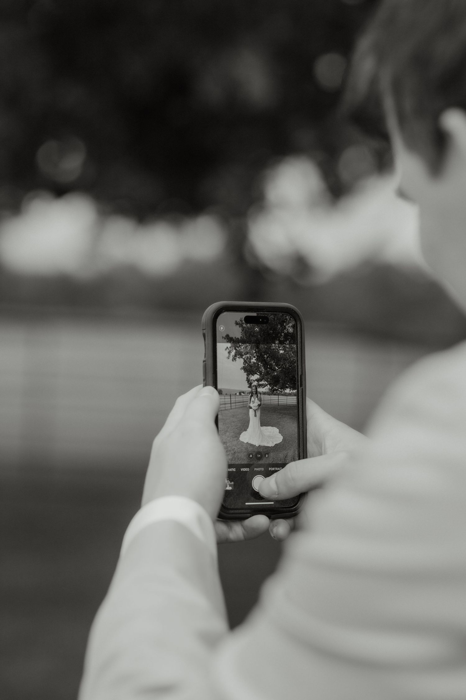 Black and white photo of groom photographing bride in wedding dress on phone at outdoor wedding venue.