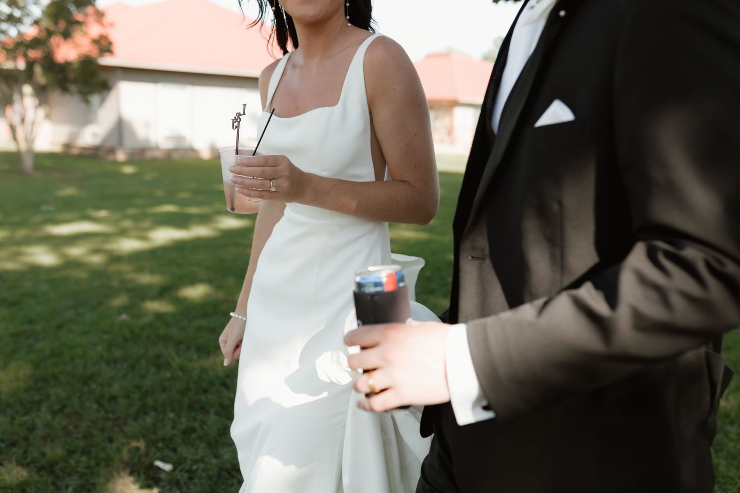 Bride in white dress and groom in black suit holding drinks outdoors at wedding.