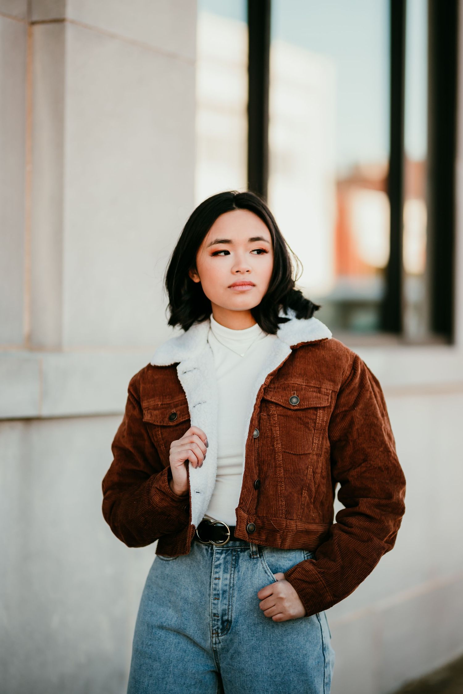 Young Asian woman in brown corduroy sherpa jacket and light blue jeans posing outdoors against a white wall.