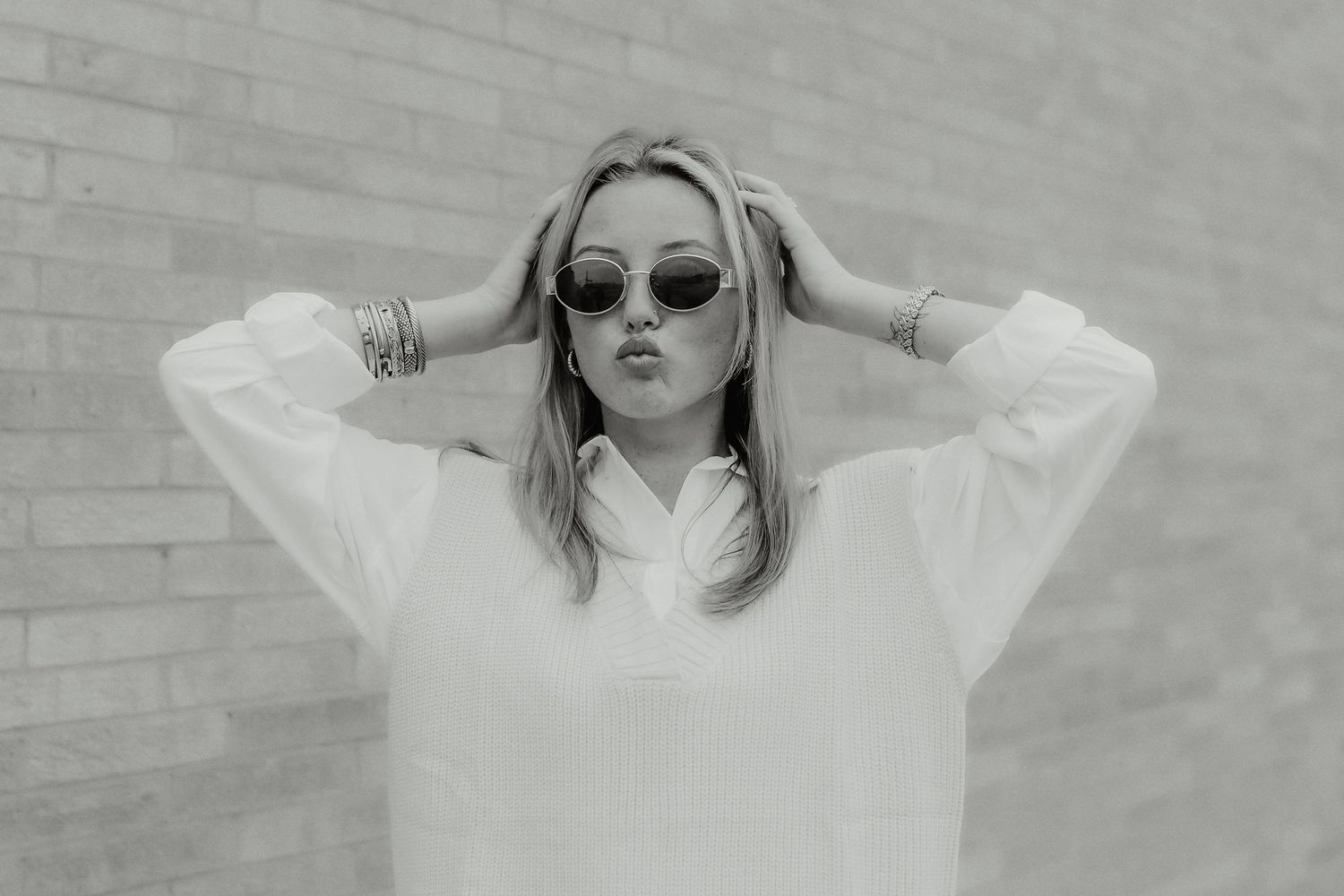 Black and white photo of blonde woman in white shirt and sunglasses posing by brick wall.