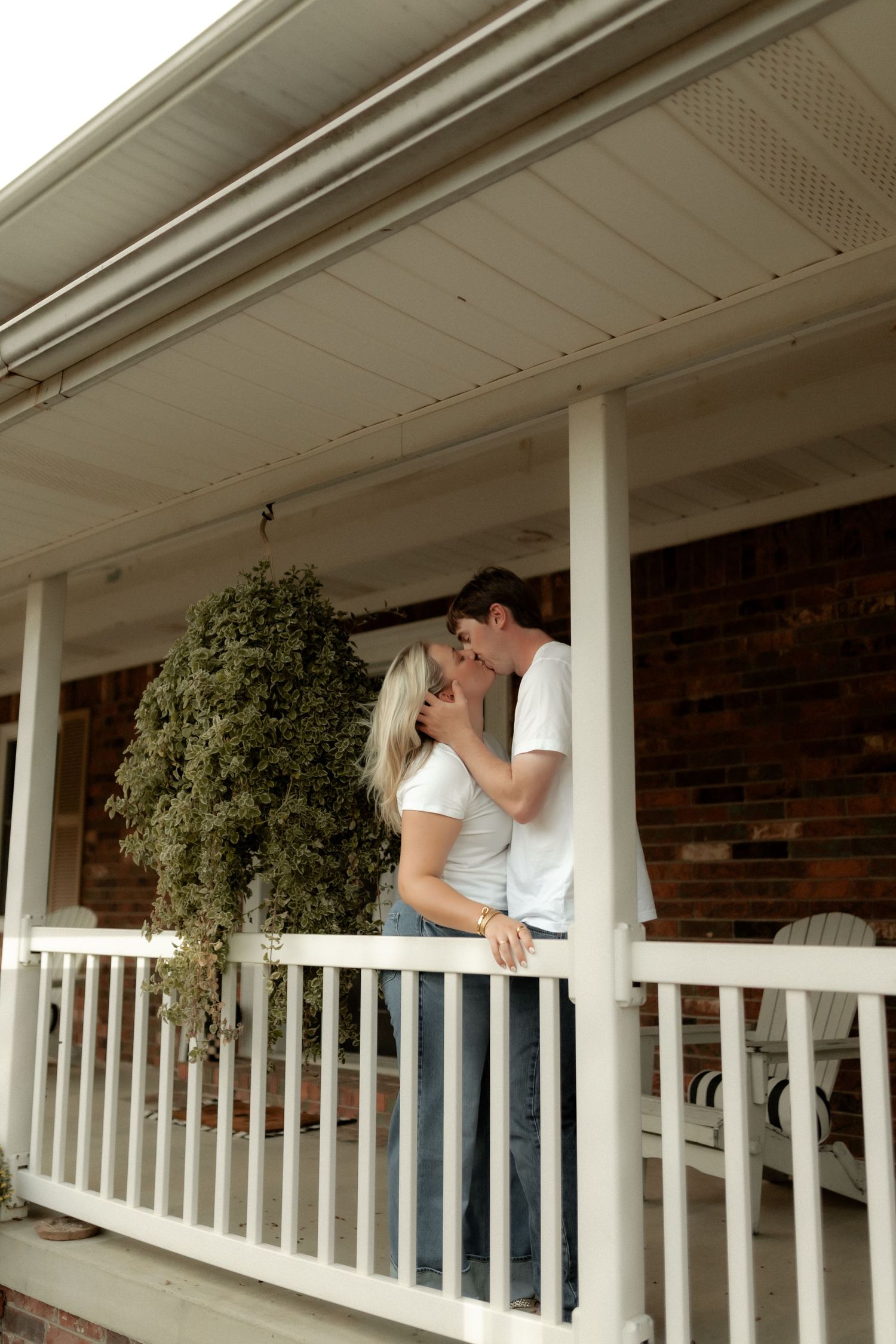 Romantic couple sharing a tender kiss on a white-railed front porch with lush hanging greenery.