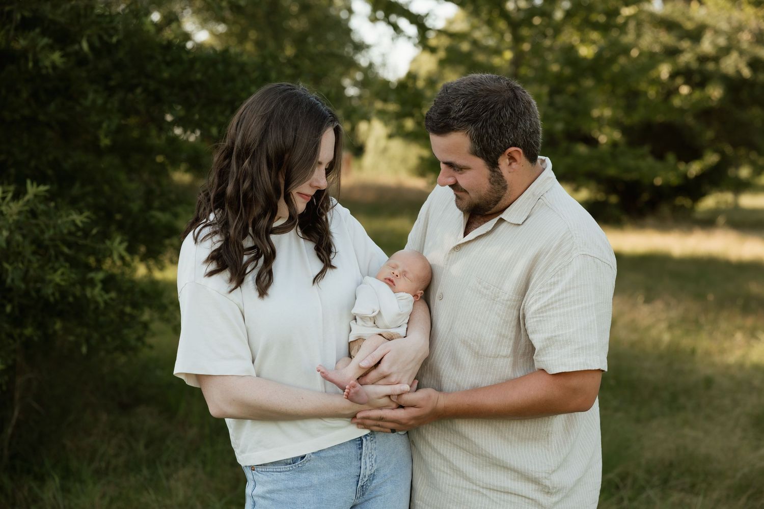 New parents smiling down at their newborn baby in a lush green outdoor setting.