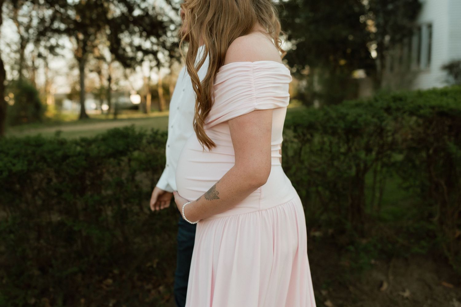 Couple embracing from behind outdoors, woman wearing a soft pink off-shoulder maternity dress in a garden setting.