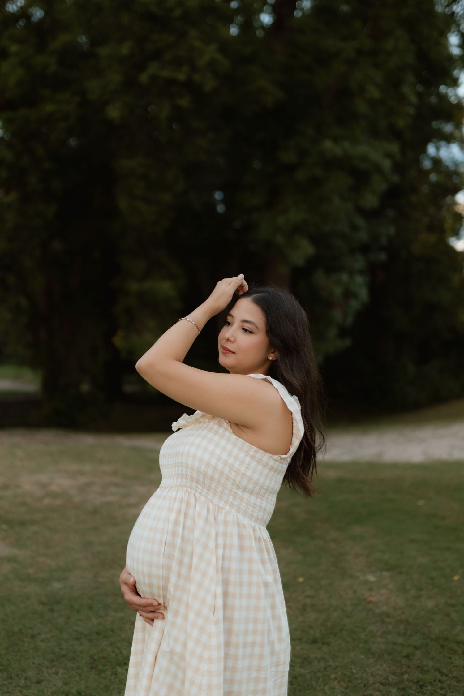 Pregnant woman in white gingham dress posing gracefully outdoors with hand in hair near trees.