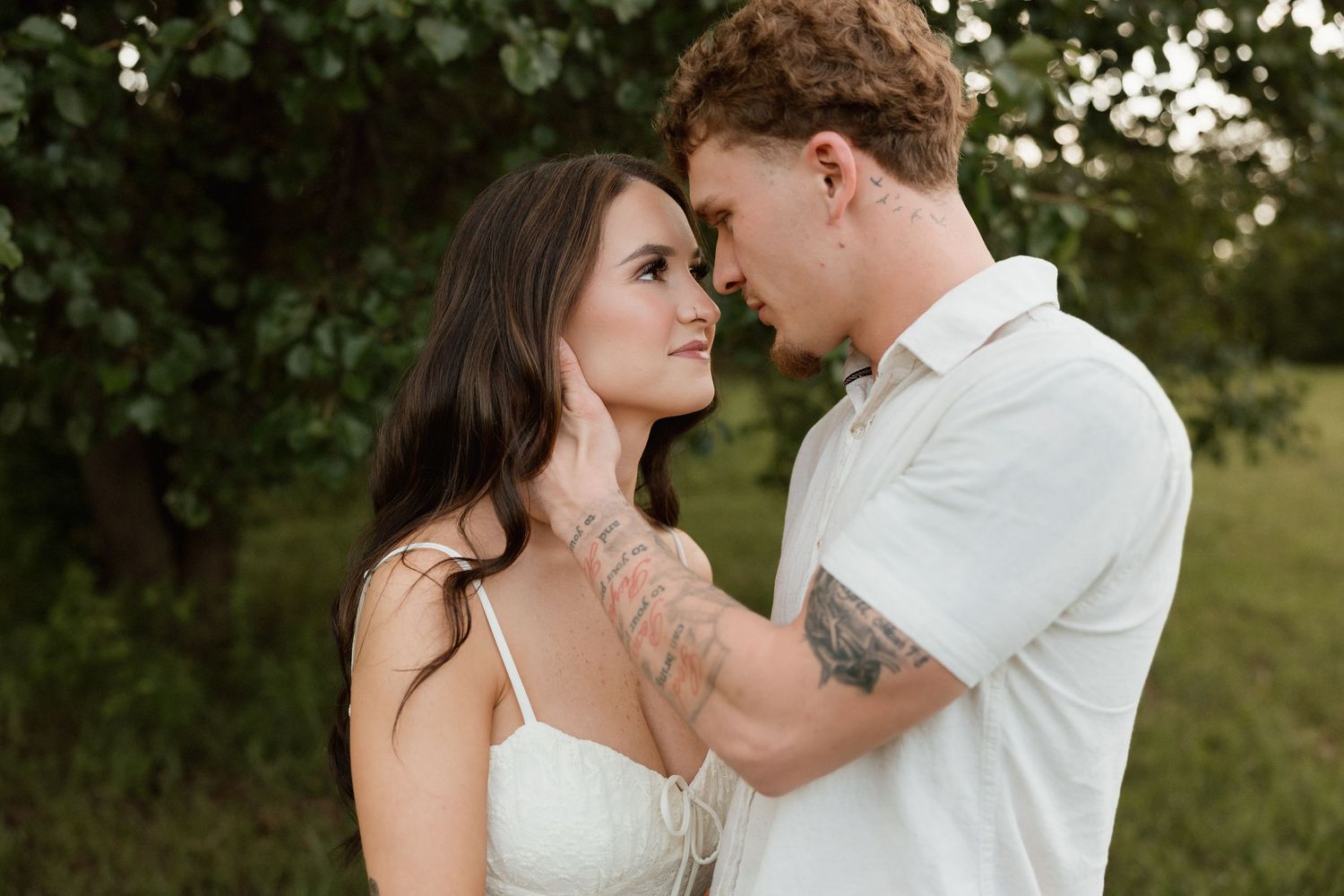 A couple with tattoos share an intimate moment touching foreheads in a green outdoor setting.