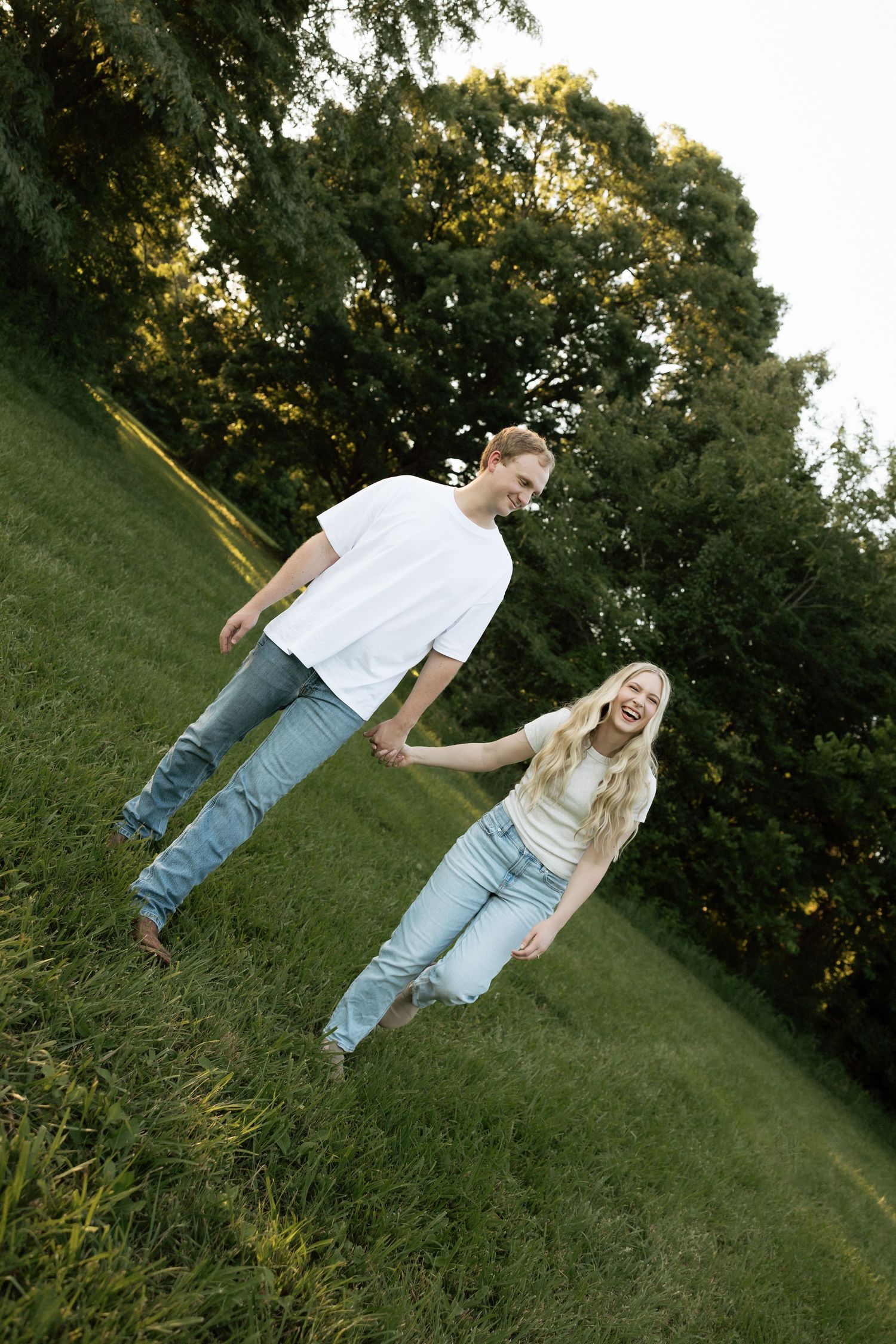 A joyful couple holding hands and walking barefoot on a grassy hill surrounded by trees in golden hour light.