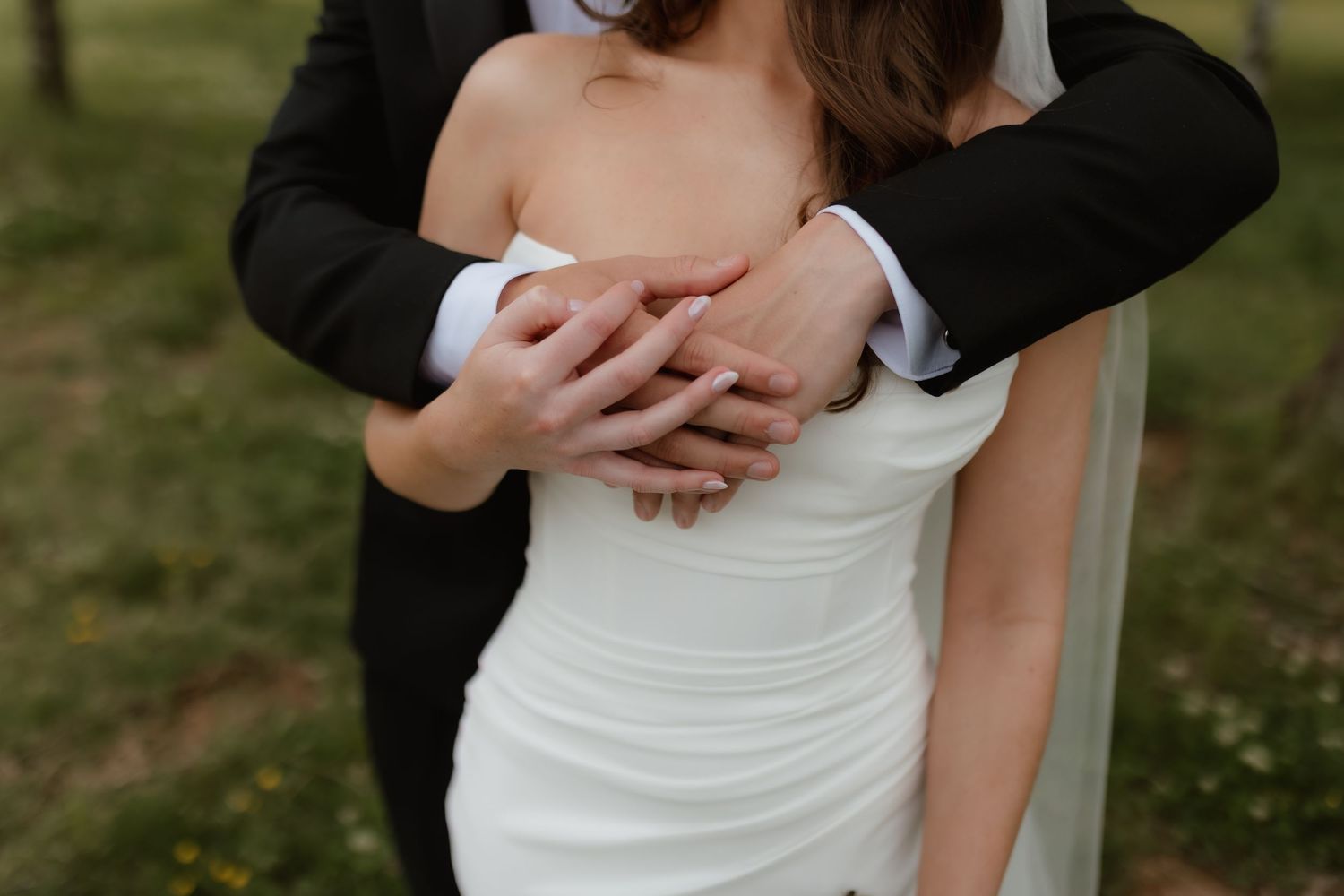 Groom embracing bride in white strapless wedding gown from behind outdoors.