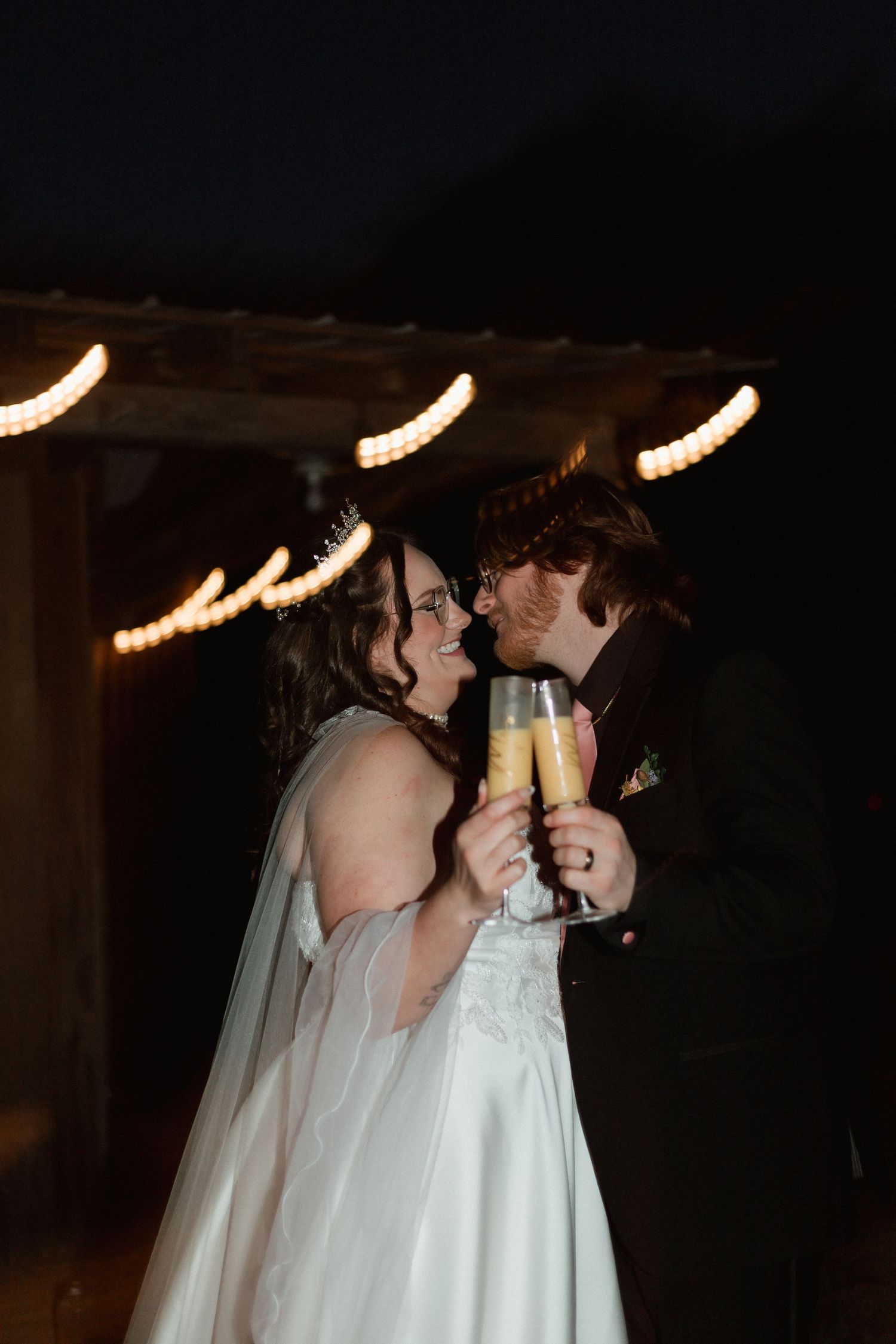 Smiling newlyweds toasting with champagne flutes at their outdoor evening wedding reception.