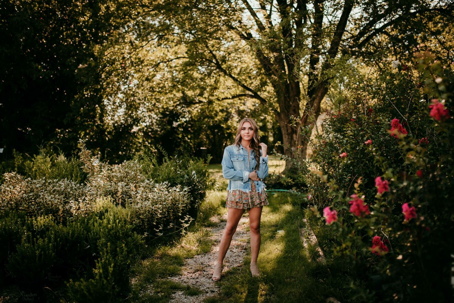 Young woman in denim jacket and plaid shorts stands on garden path surrounded by colorful flowers.