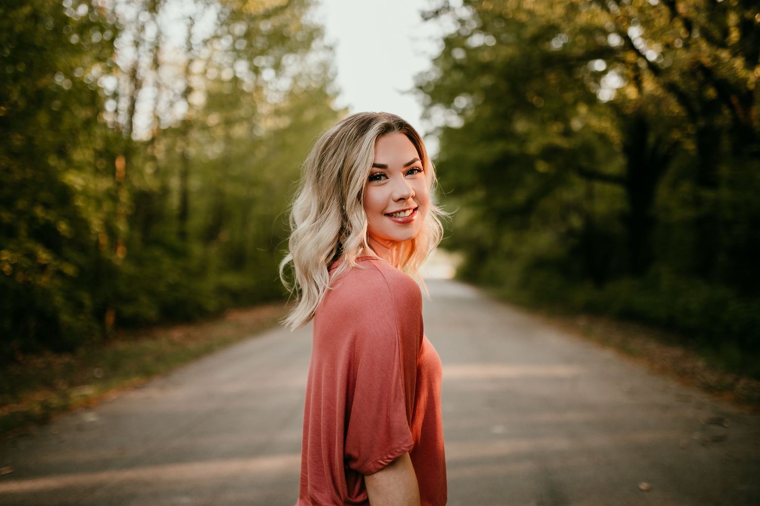 Smiling blonde woman in rust top glancing back on tree-lined road at golden hour.