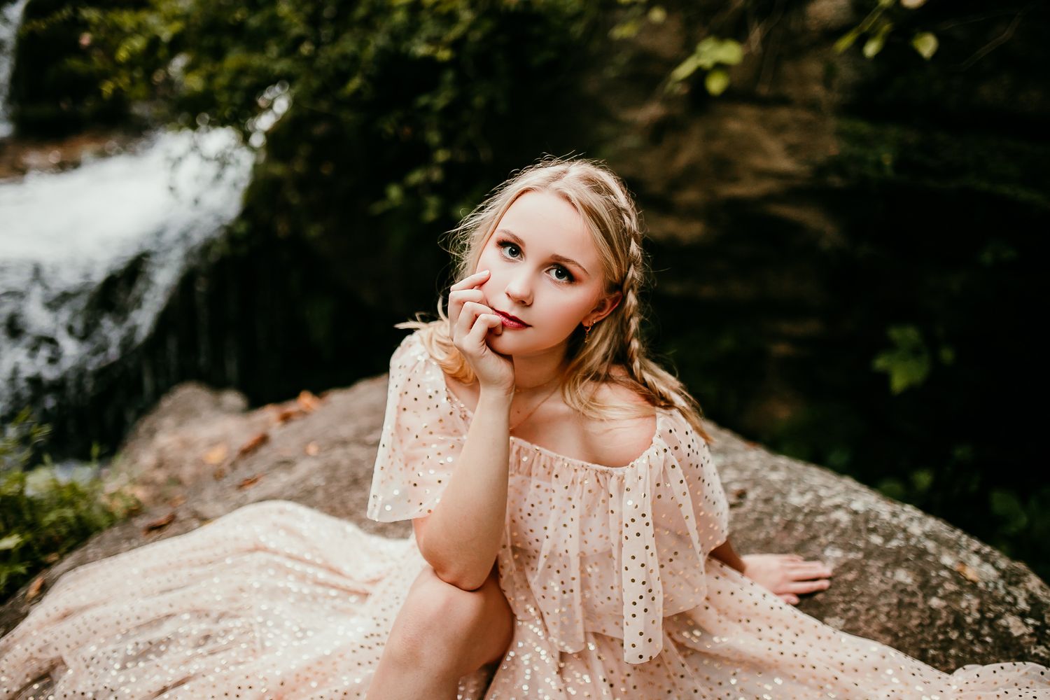 Young blonde girl in off-shoulder blush floral dress sits on rock near waterfall, gazing upward.