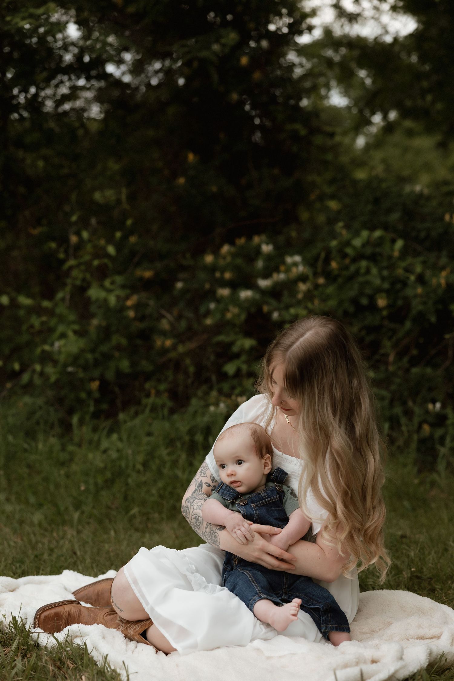 Mother with long blonde hair sitting on blanket holding baby outdoors.