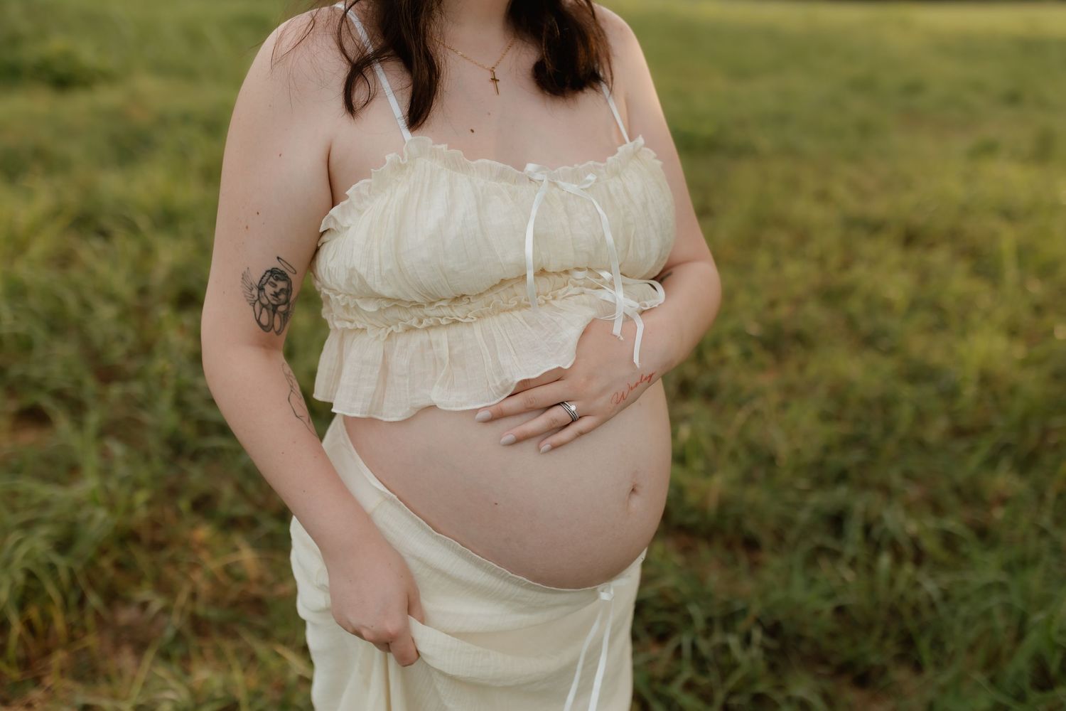 Close-up of pregnant womans bare baby bump in cream two-piece outfit in grassy field.