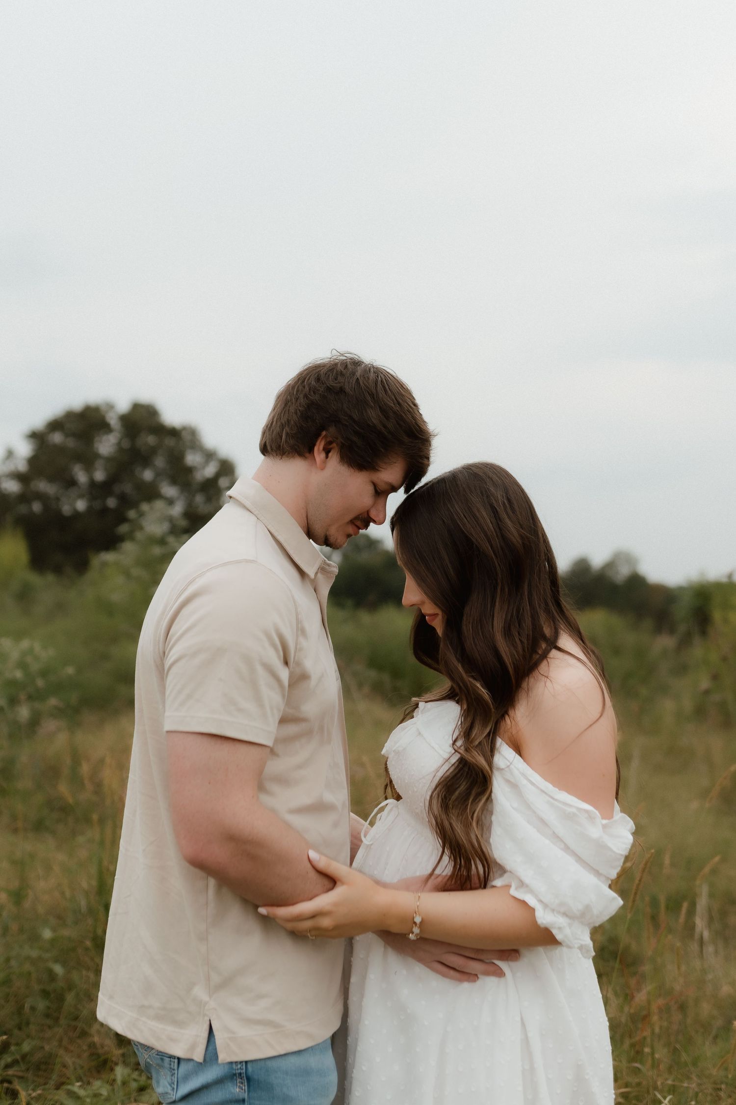 Pregnant couple stands forehead-to-forehead in a green meadow sharing an intimate moment.