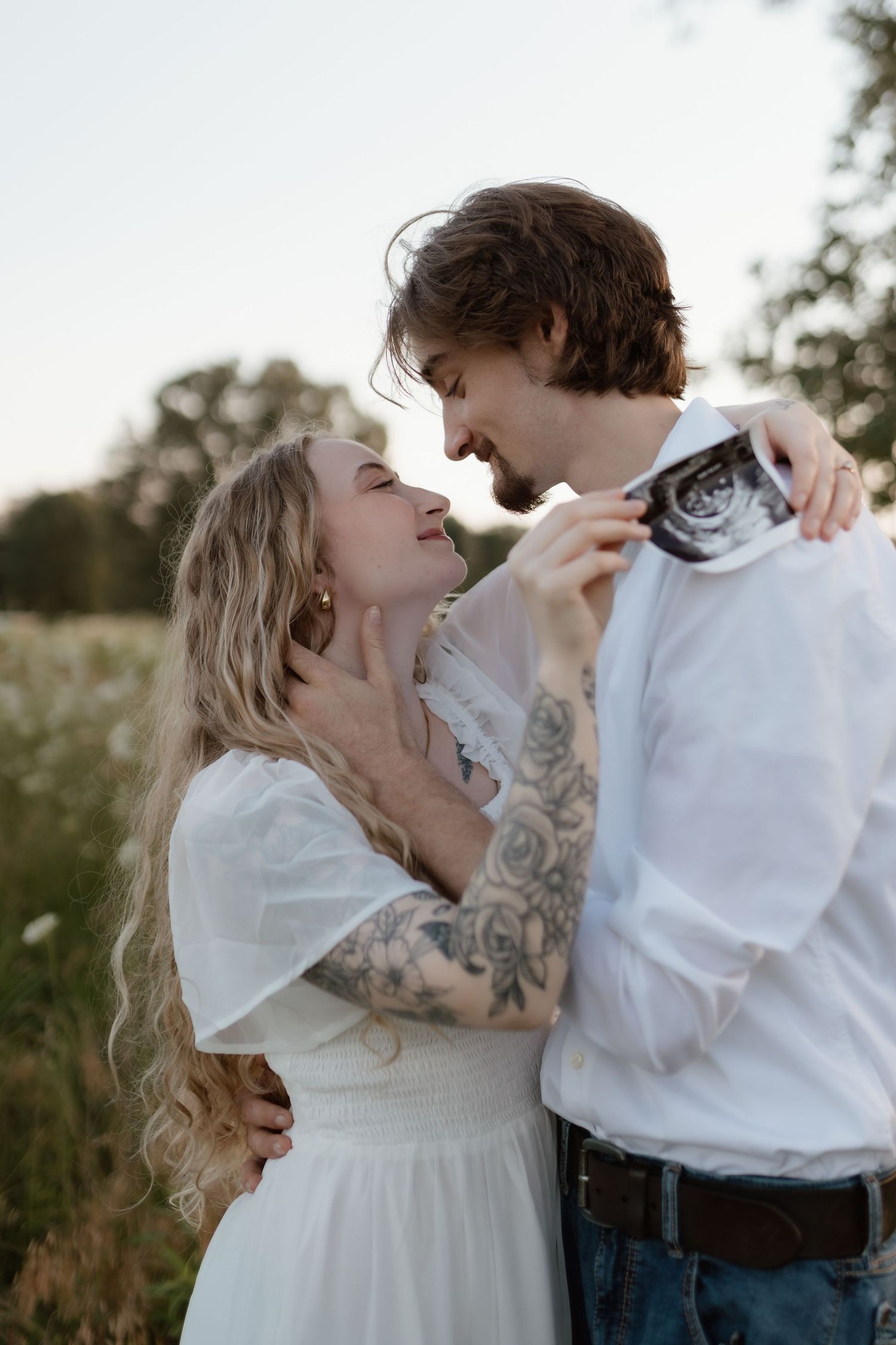 Tattooed blonde woman in white dress embracing bearded man holding ultrasound photo in a sunlit wildflower field.