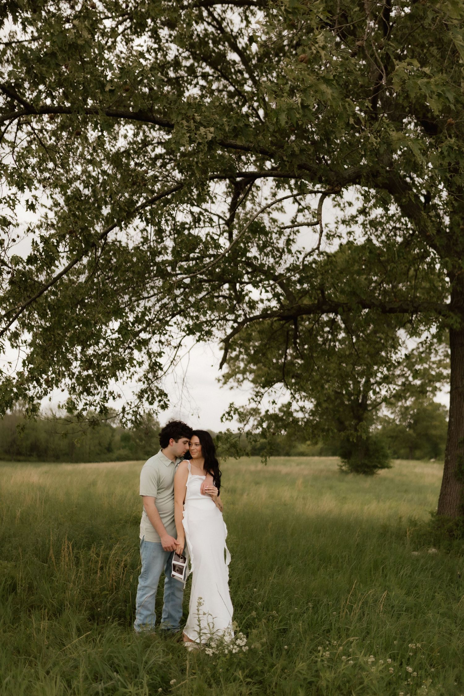 Expecting couple embracing tenderly under large tree in green meadow, woman in elegant white maternity dress.