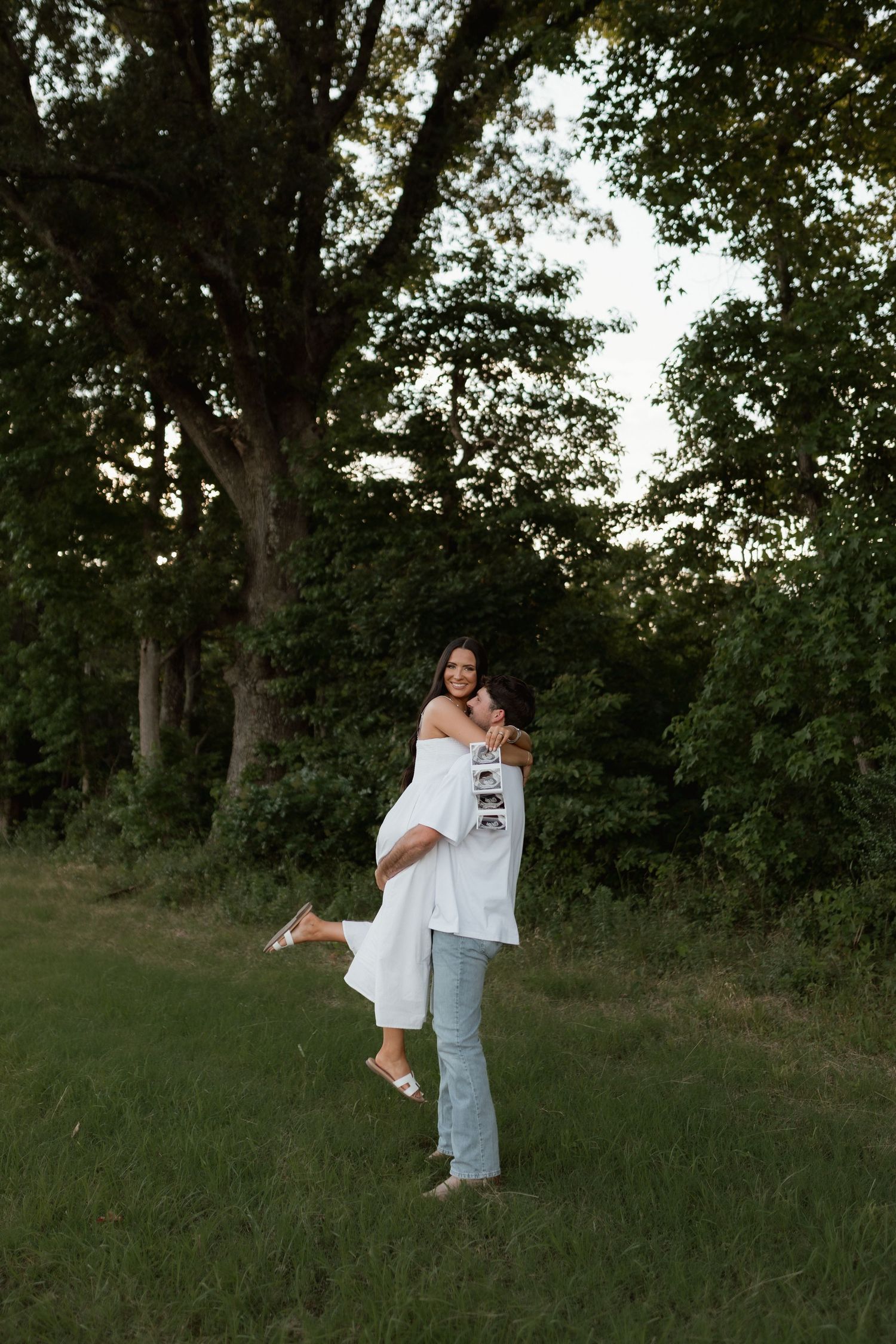 Man lifting smiling pregnant woman in white dress while holding ultrasound photos in a green field surrounded by trees.