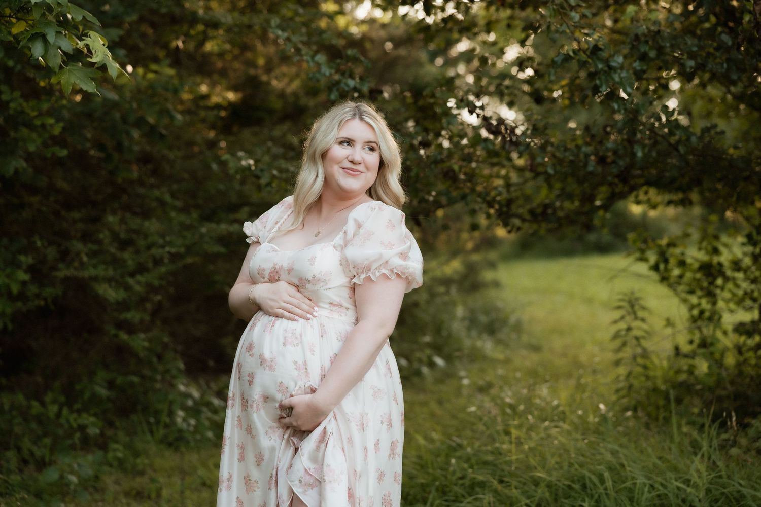 Smiling pregnant woman in a floral off-shoulder maternity dress cradling her belly in a garden.