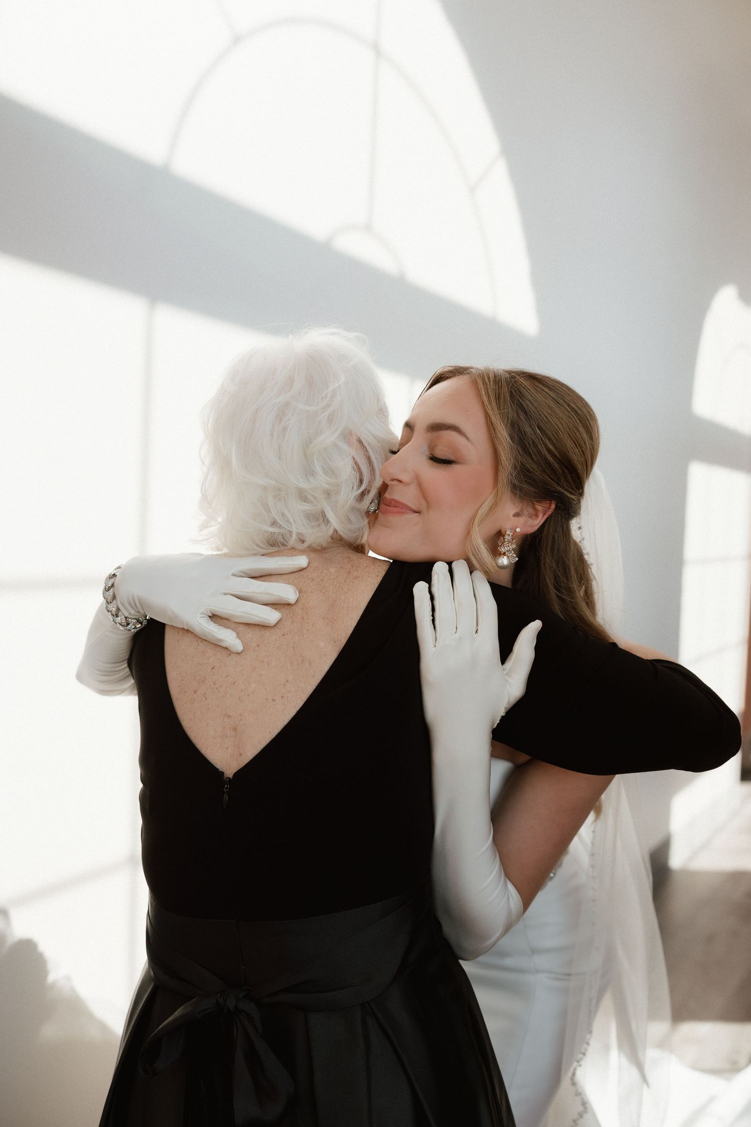 Bride in white gloves shares a tender embrace with an elderly woman in black dress.