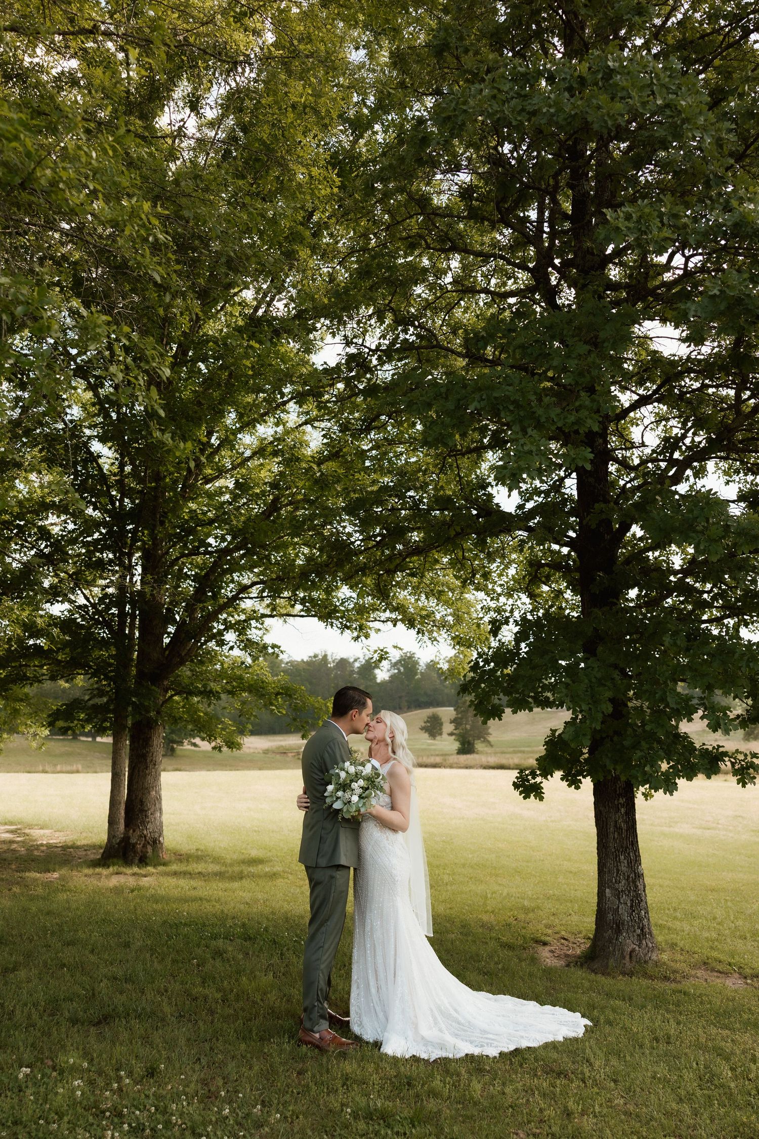 Bride and groom sharing romantic kiss under tall oak trees in lush green countryside setting.
