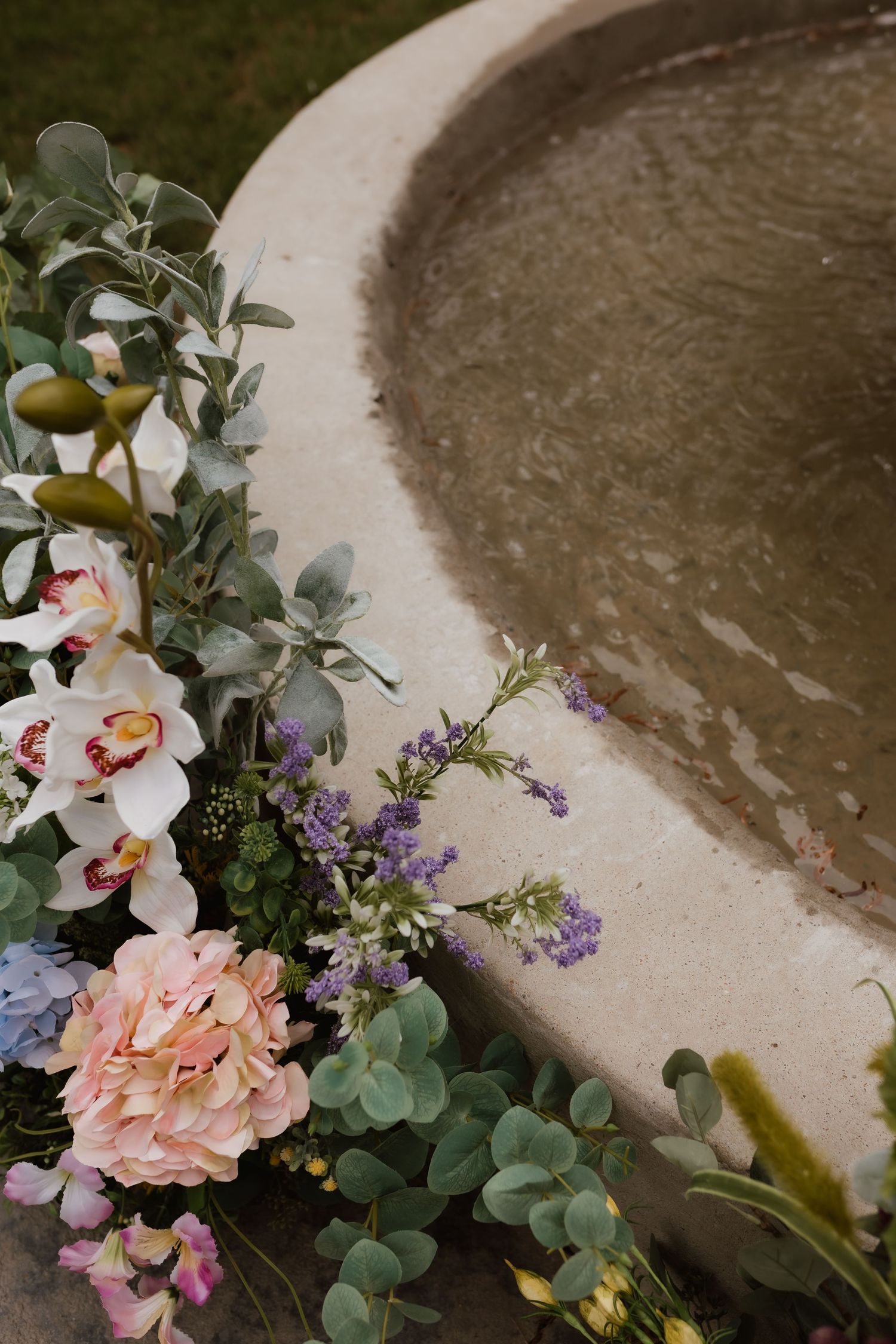 Elegant floral arrangement with roses, orchids, lavender, and eucalyptus beside a stone fountain.