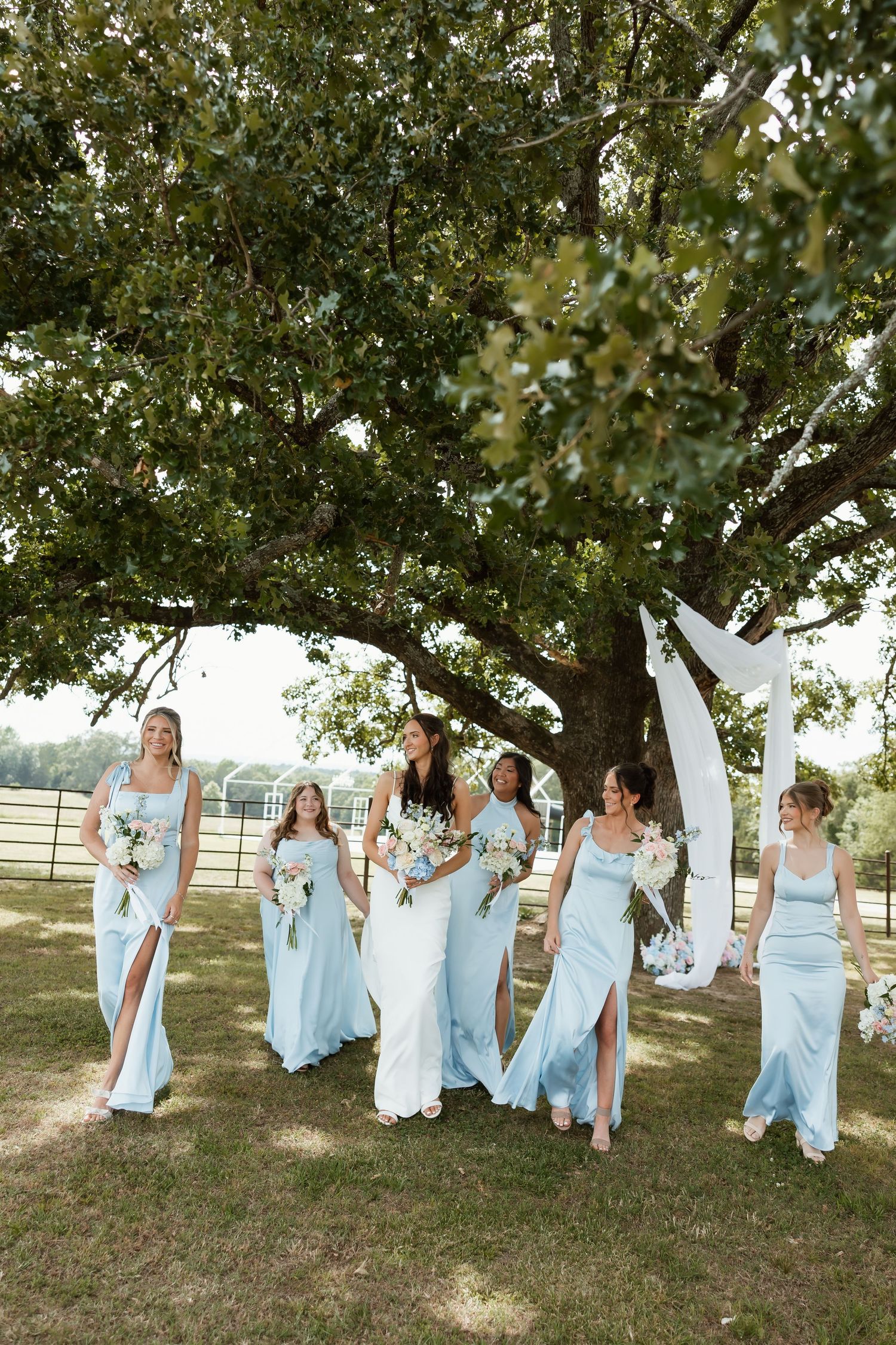 Bridesmaids in light blue satin dresses walking with bride under large oak tree outdoors.