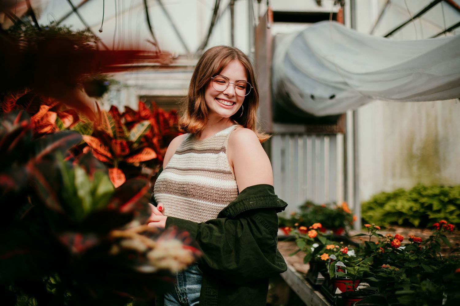 Happy teenage girl with glasses smiles among vibrant colorful flowers inside a sunlit greenhouse.