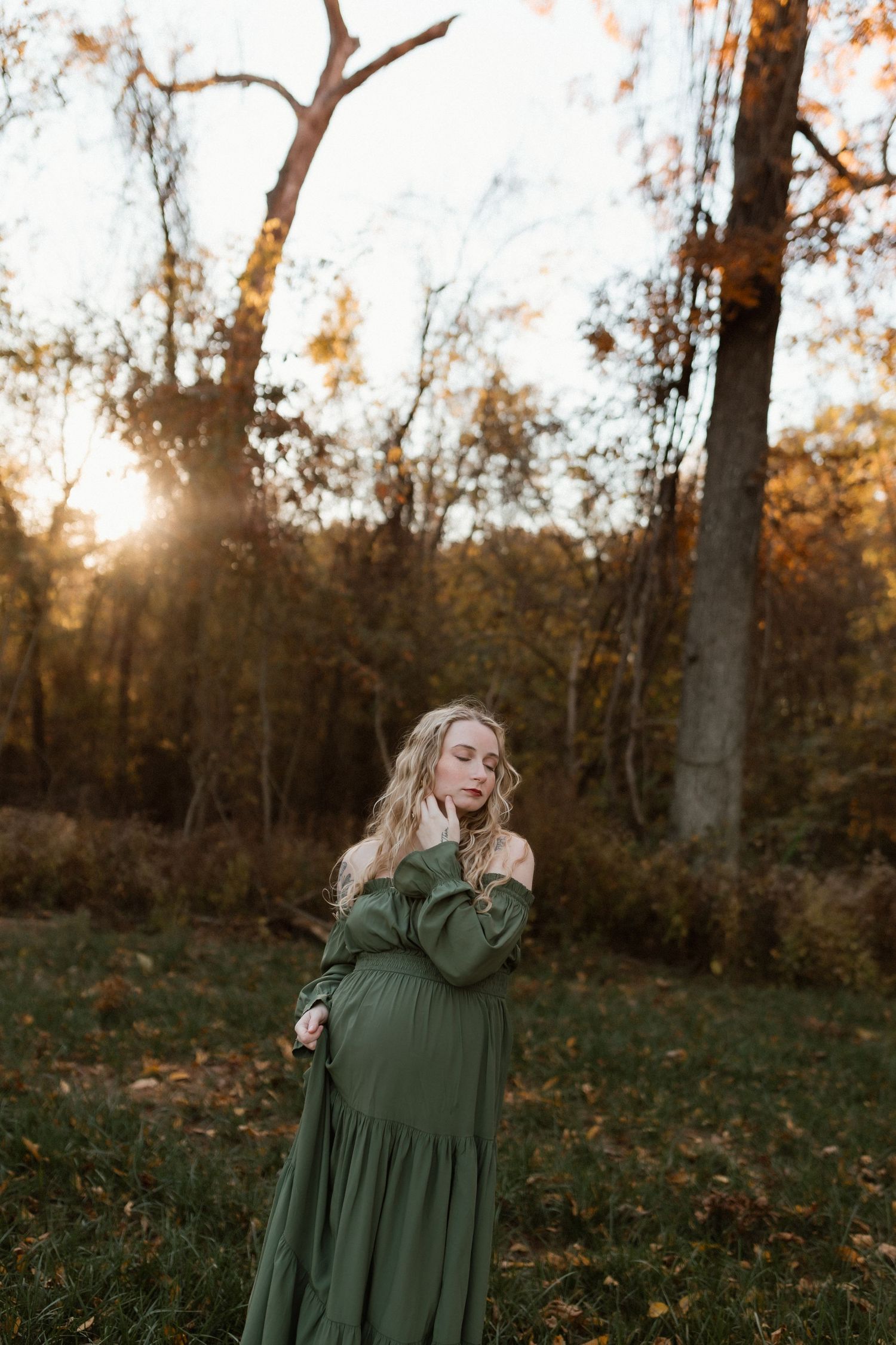 Pregnant woman in green off-shoulder dress poses in autumn forest at golden hour sunset.