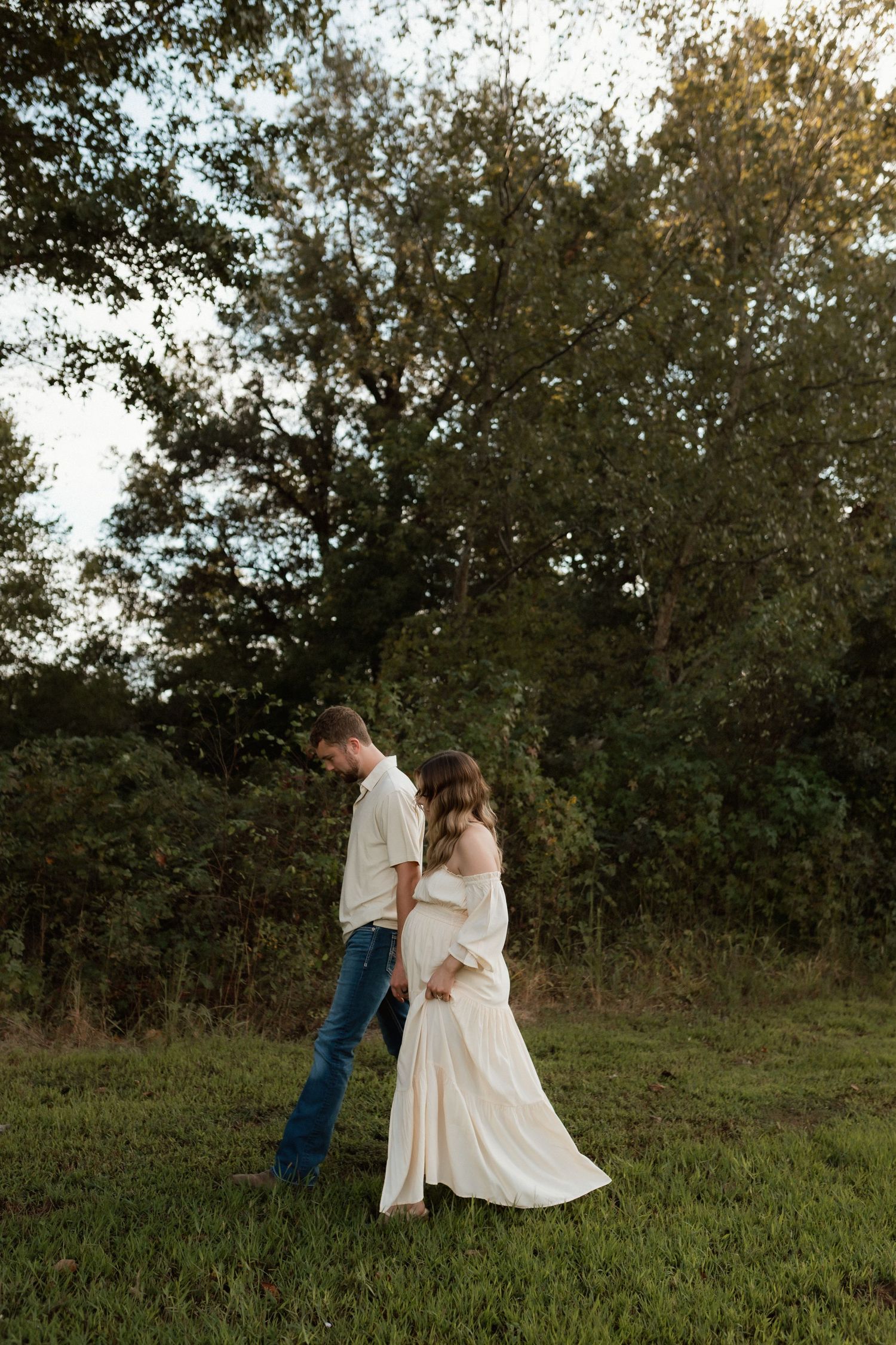 Pregnant couple walking hand-in-hand through green field during golden hour maternity shoot.