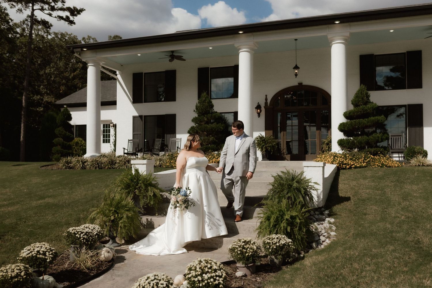 Bride and groom walk hand in hand in front of a grand white estate on their wedding day.