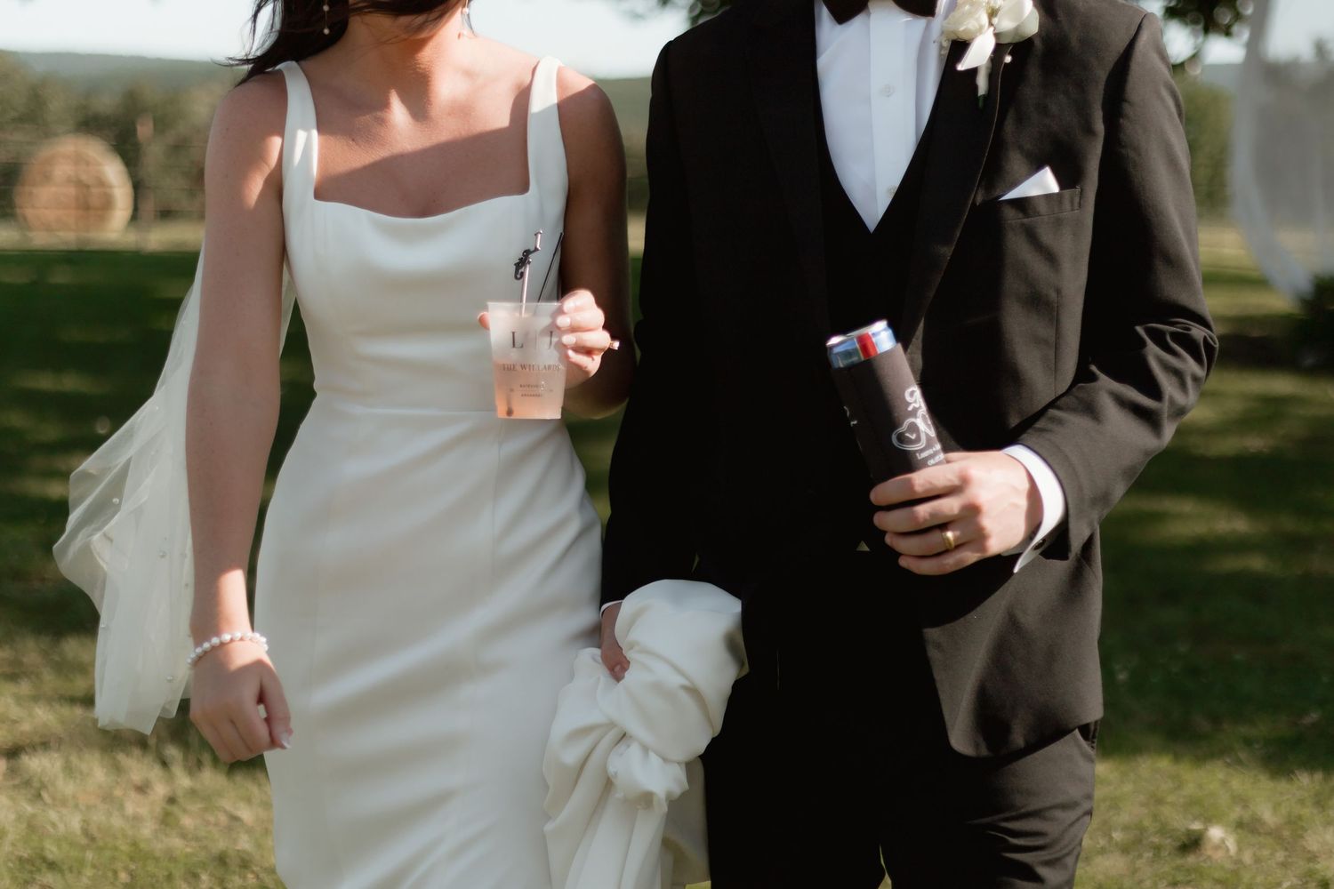 Bride in sleek white gown and groom in black tuxedo walk together holding drinks outdoors.