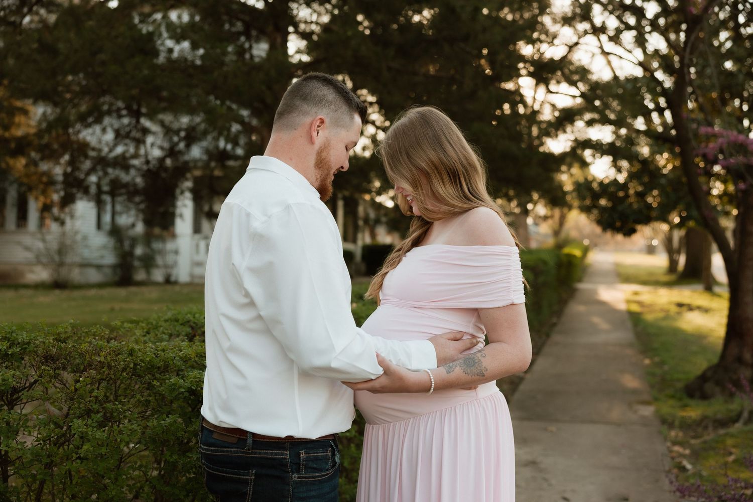 Couple sharing a tender moment on a garden path, woman in a blush off-shoulder maternity gown at golden hour.