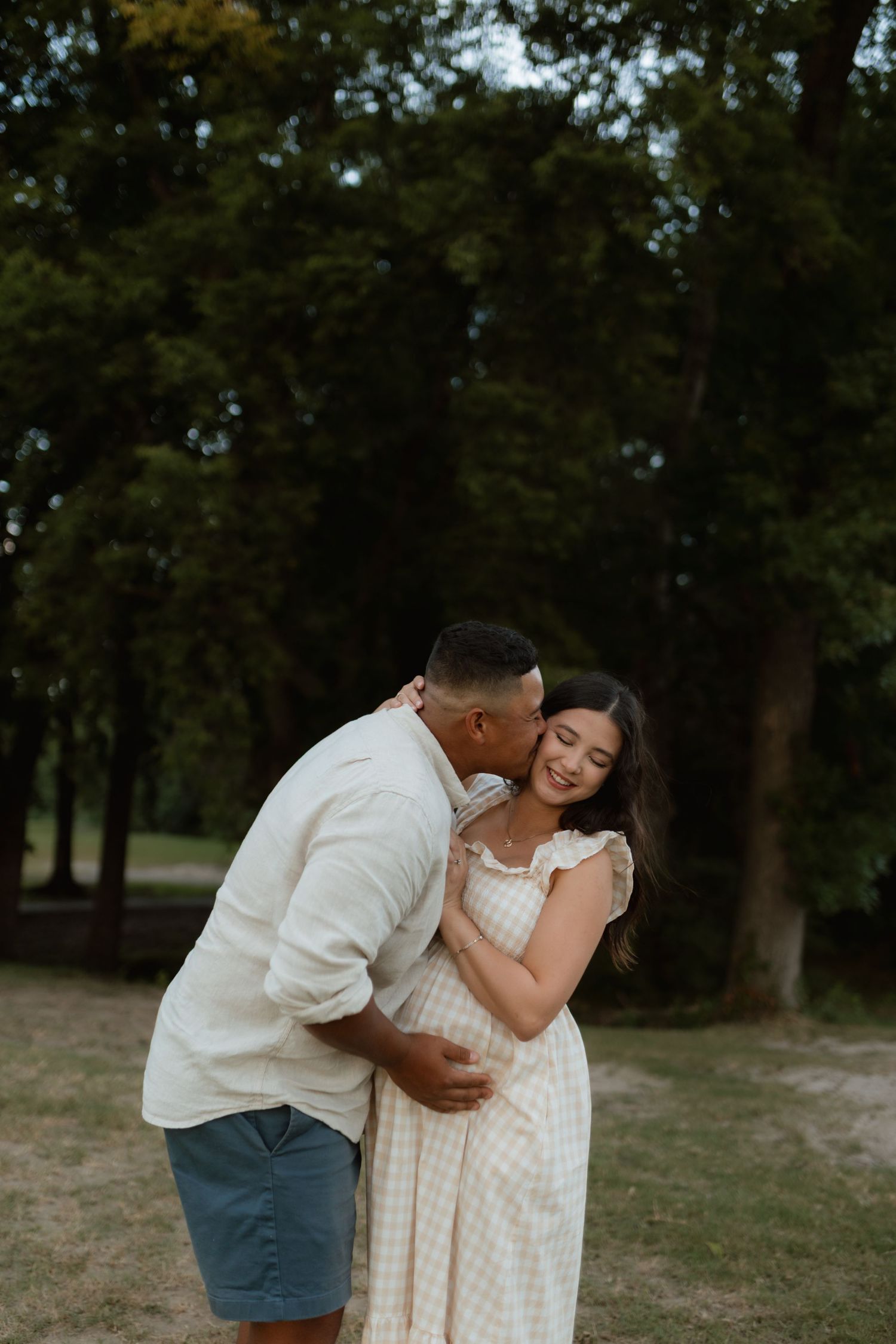 Man kissing pregnant womans cheek while embracing her from behind near tall trees outdoors.