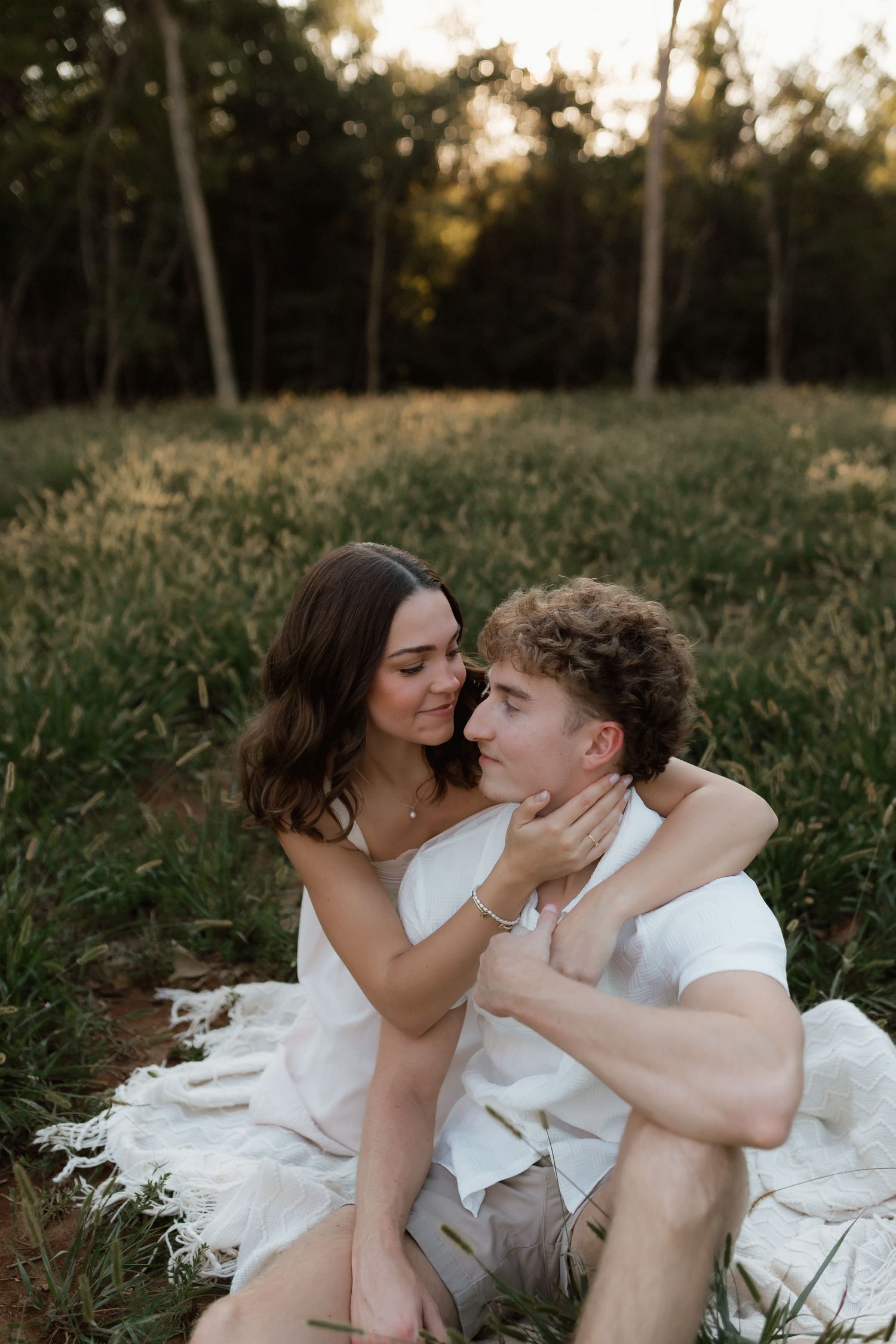 Couple sitting on white blanket in meadow at dusk, woman gently holding mans face romantically.