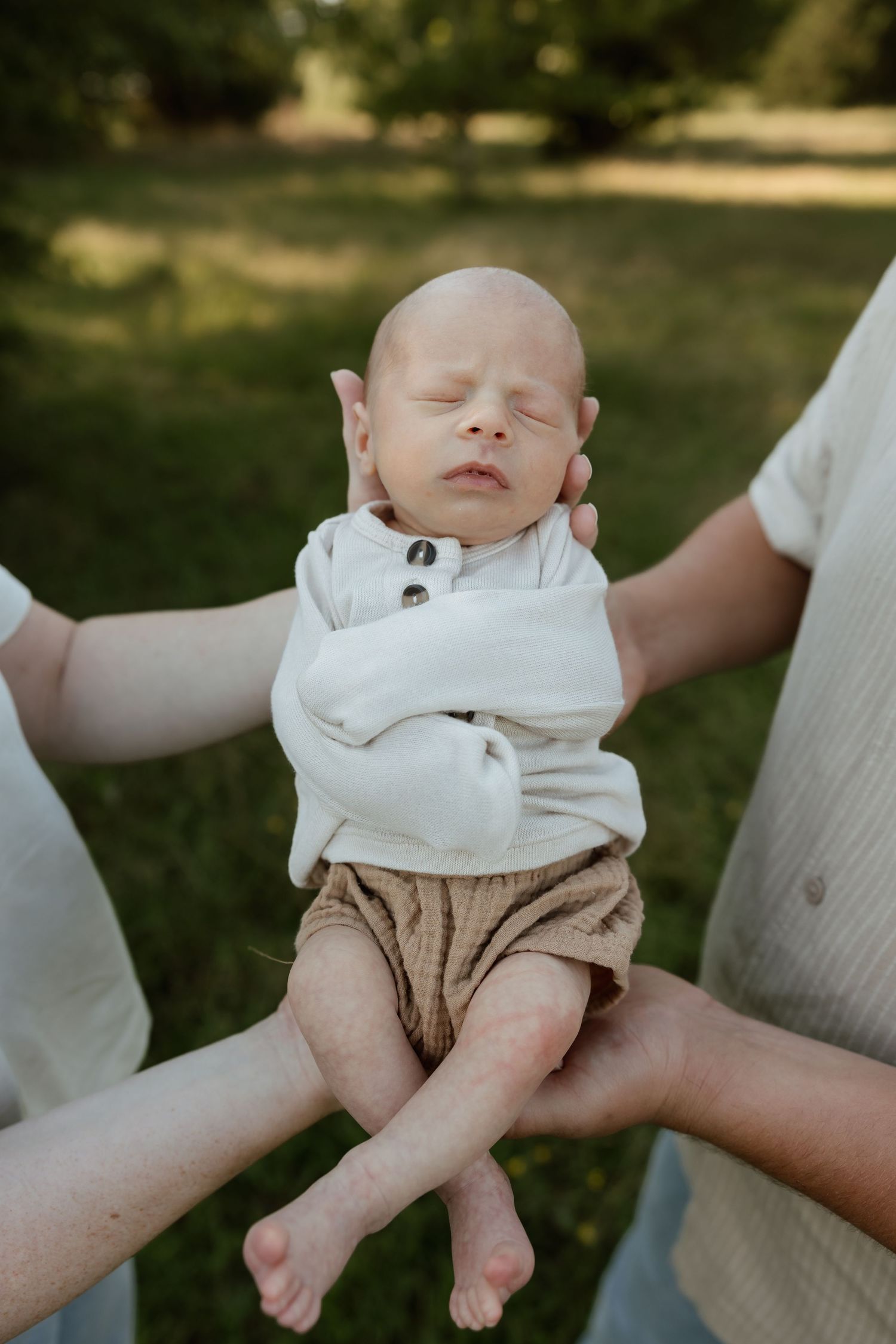 Newborn baby in white onesie sleeping peacefully while being held by parents outdoors.