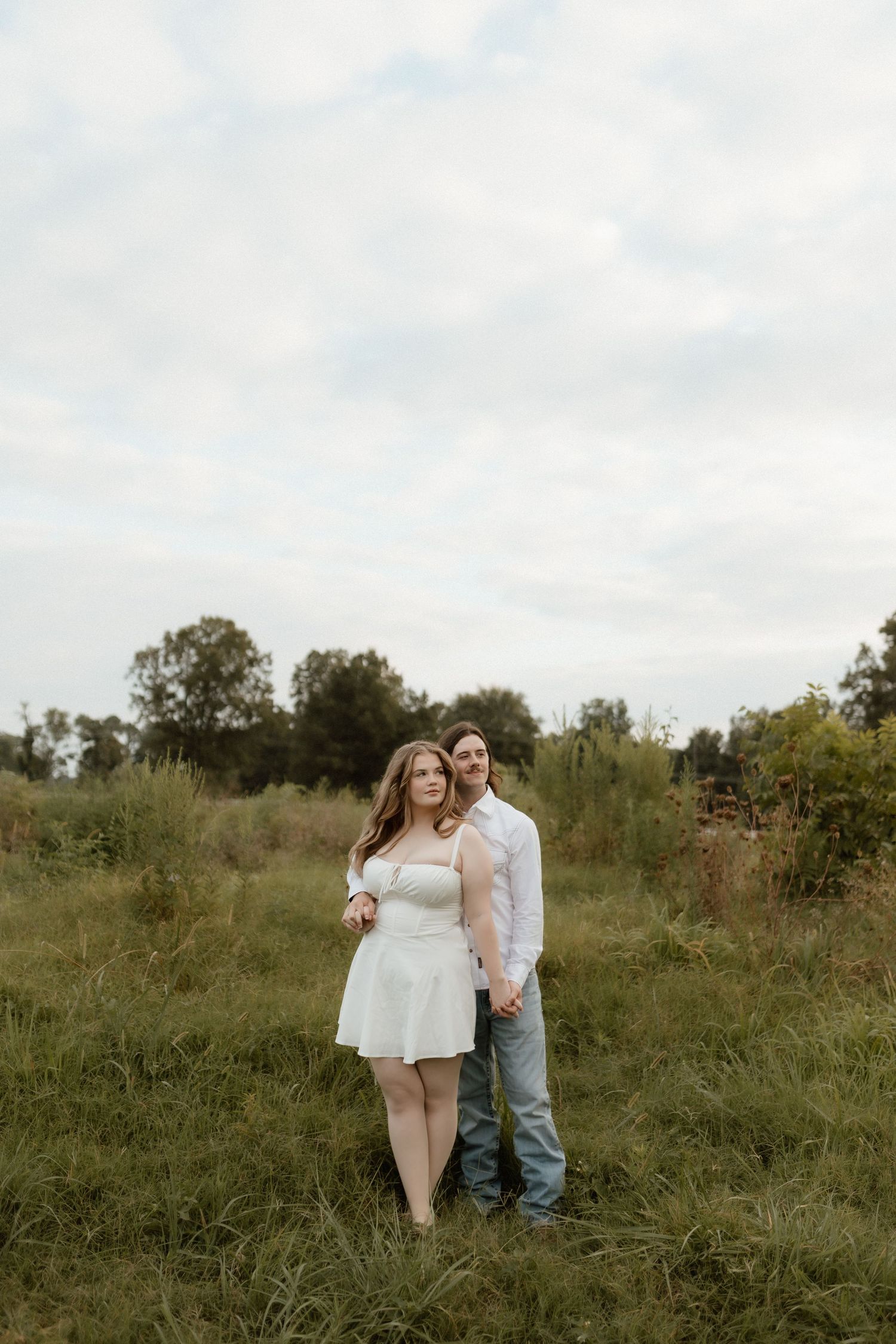Couple standing together in lush green field under cloudy sky, woman in white mini dress, man in white shirt.