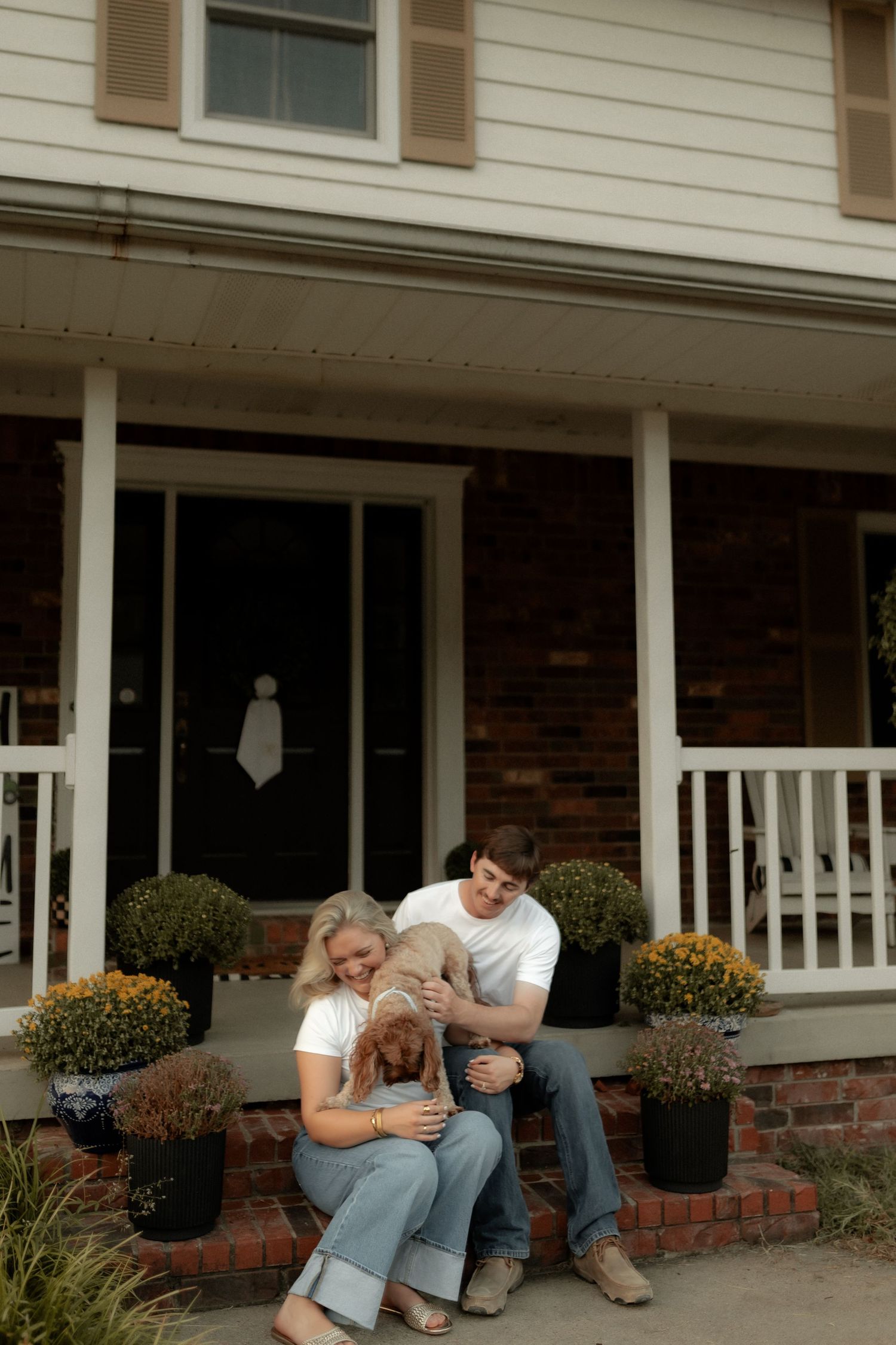 Couple sitting on brick porch steps with golden retriever puppy in front of charming brick home with white railings.