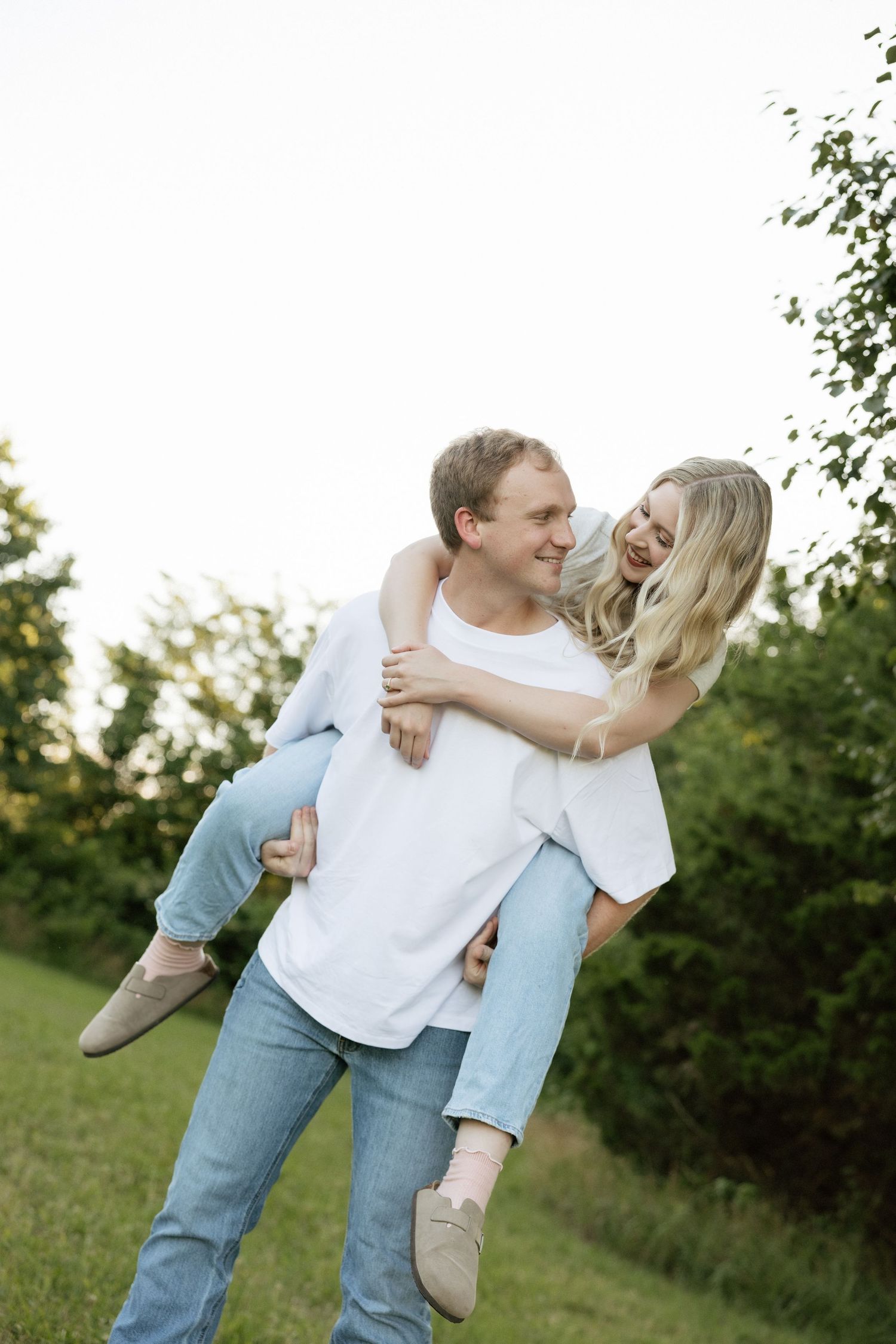 A happy couple laughing outdoors as a man gives a smiling blonde woman a piggyback ride in a lush green park.
