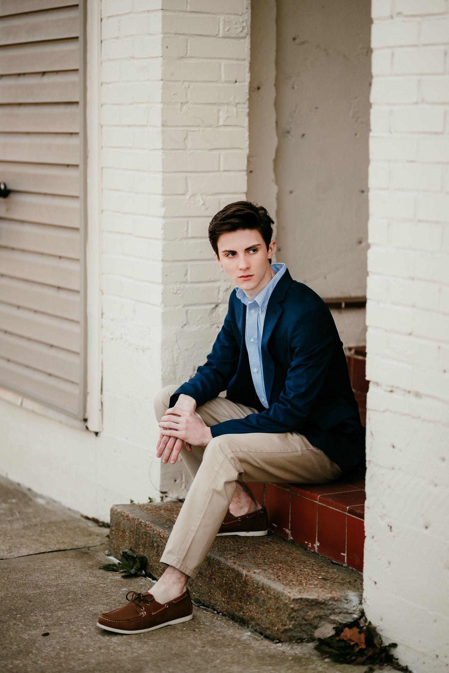 Young man in navy blazer sits on brick steps beside white painted building exterior.