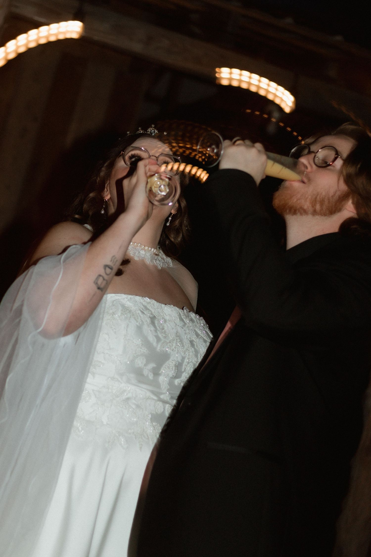 Bride and groom drinking champagne together at their wedding reception under warm string lights.
