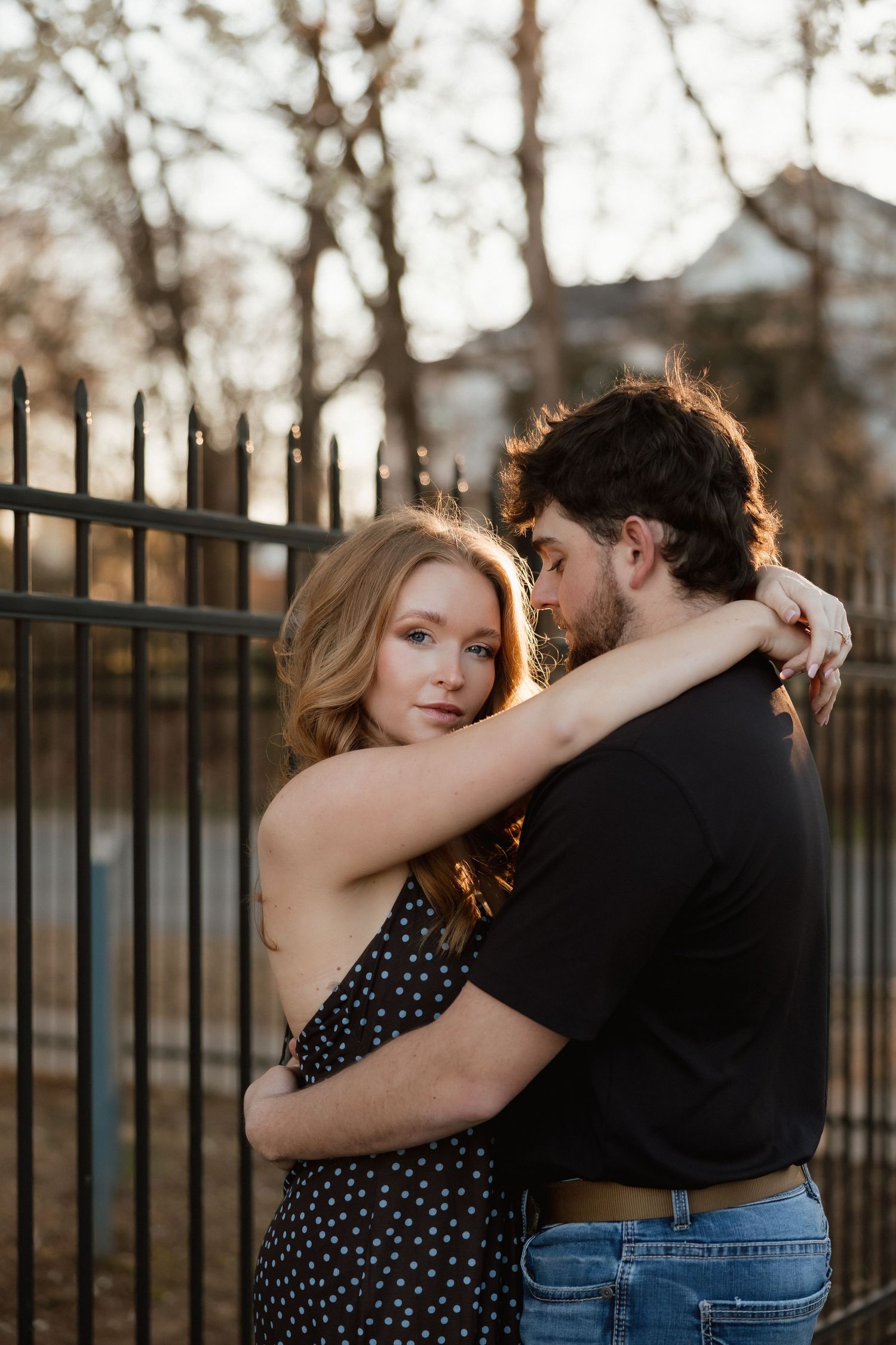 A couple embraces tenderly near a black iron fence outdoors at golden hour, woman in polka dot dress gazing at camera.