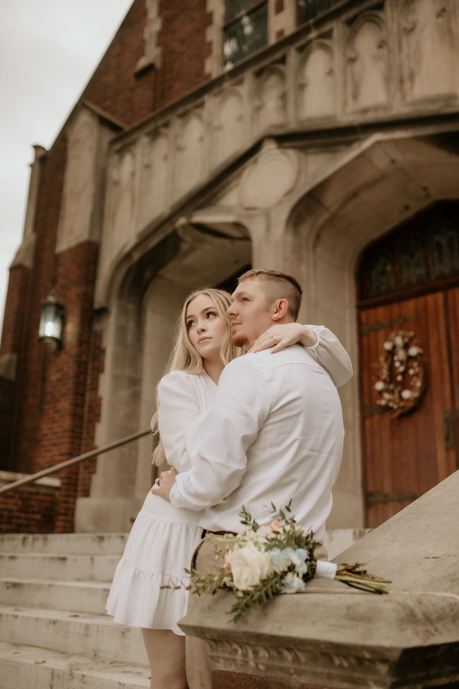 A couple embraces on church steps holding a white bouquet before a brick Gothic facade.