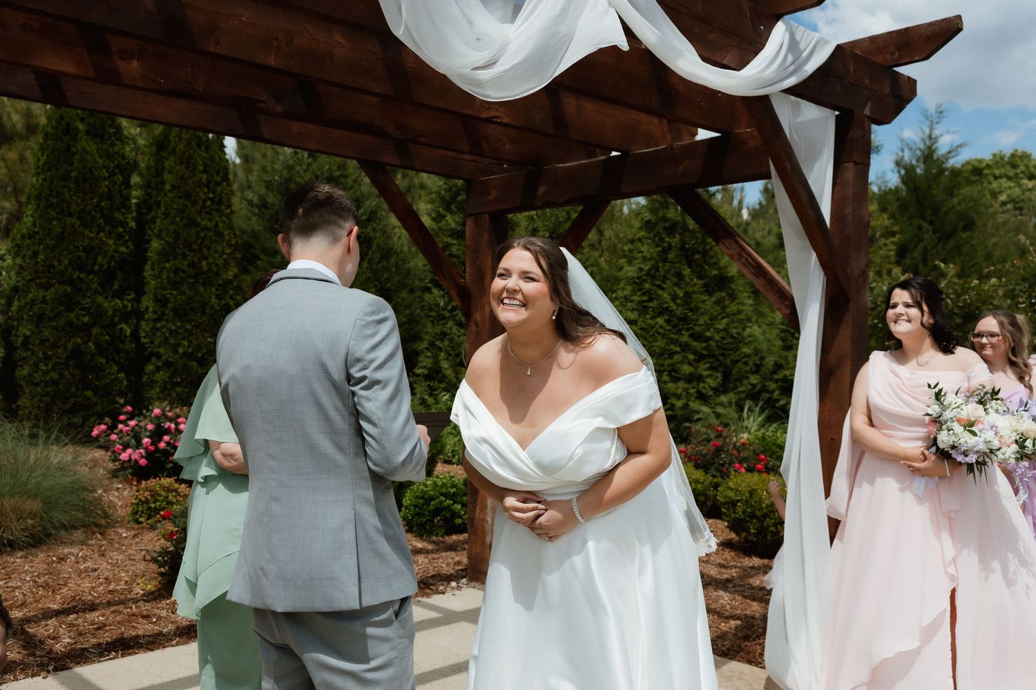 Bride smiling joyfully at groom during outdoor wedding ceremony under wooden pergola.