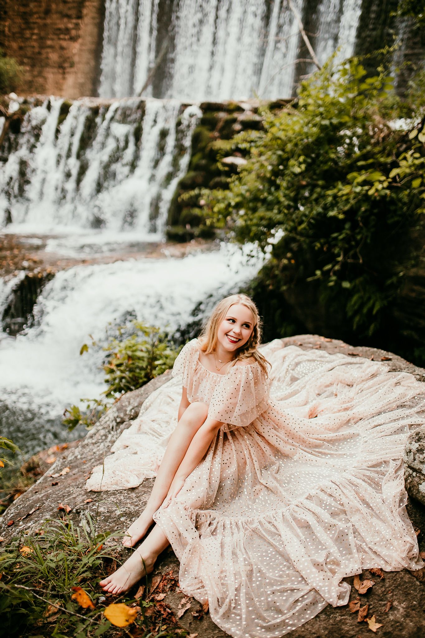 Smiling blonde woman in sparkling blush gown sits barefoot on rock with waterfall backdrop.
