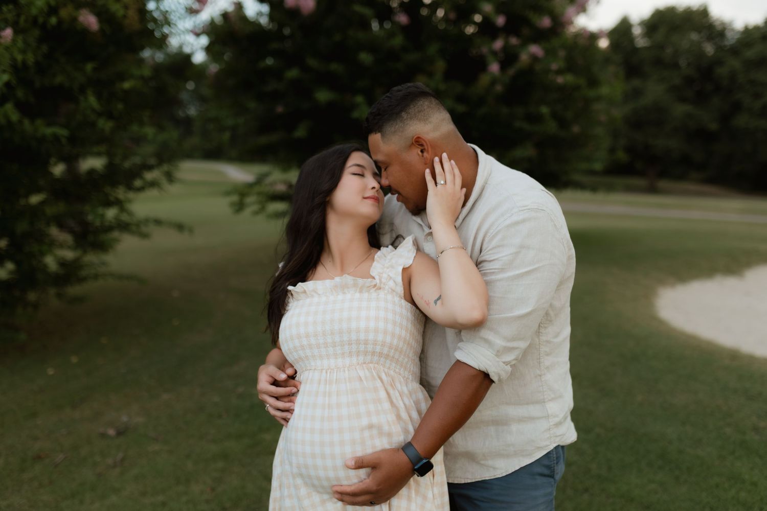 Expecting couple embracing tenderly outdoors, man kissing womans head while cradling her baby bump.