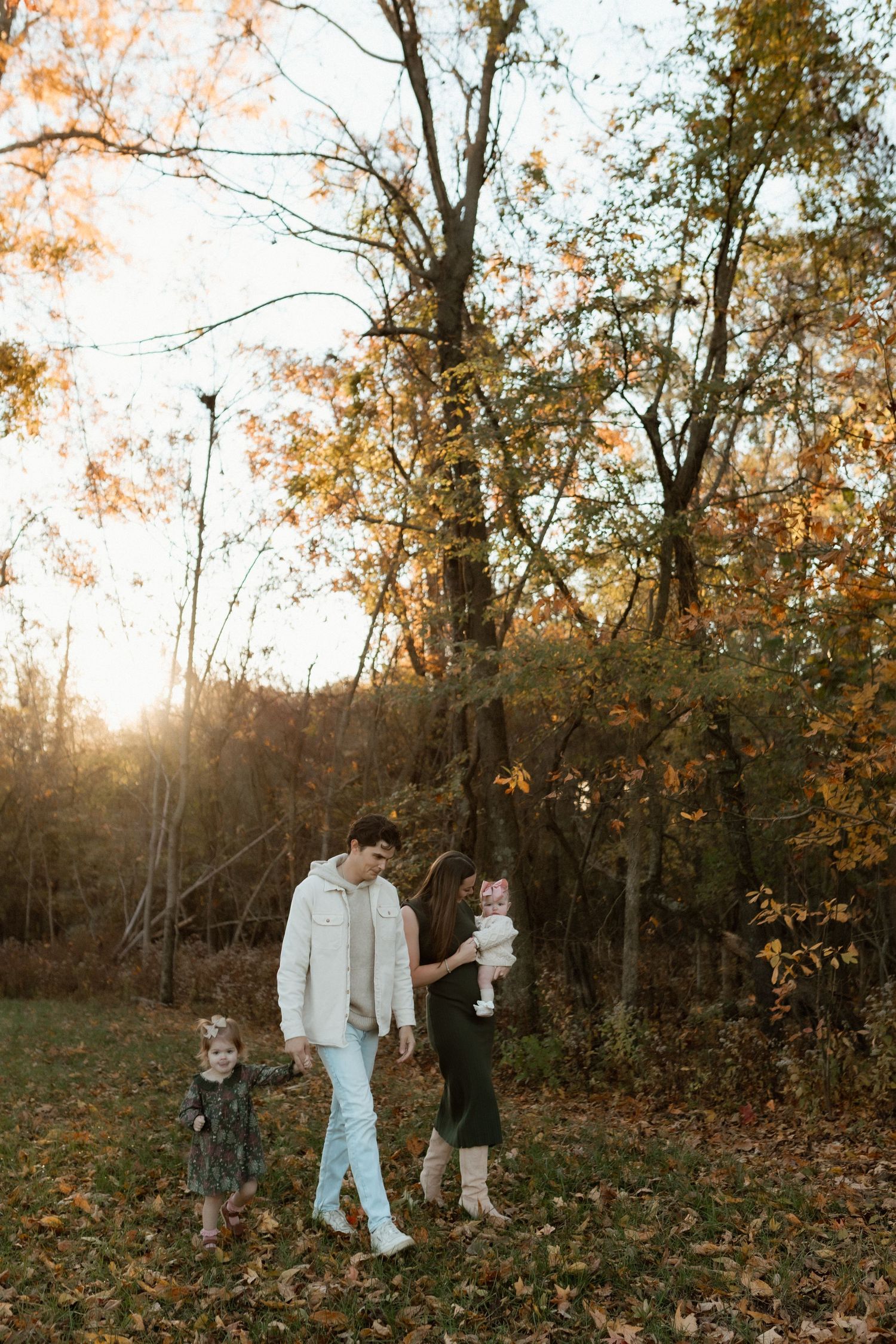 Young family of four walking together in an autumn park during golden sunset among fallen leaves.