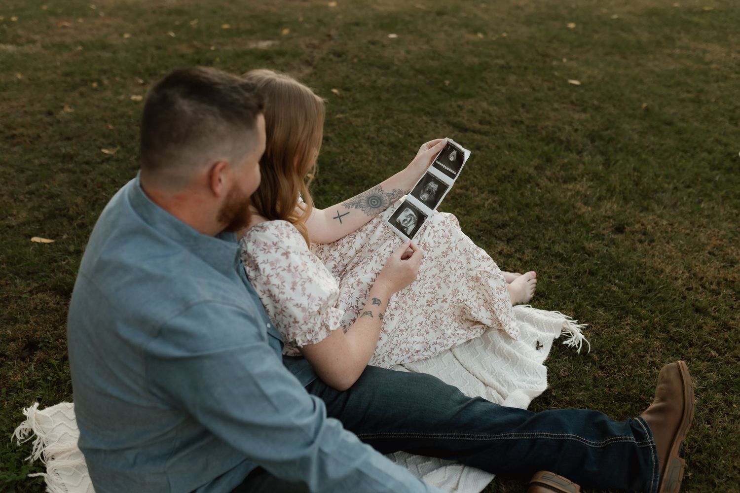 Couple sitting on blanket in grass reviewing ultrasound photos together outdoors.