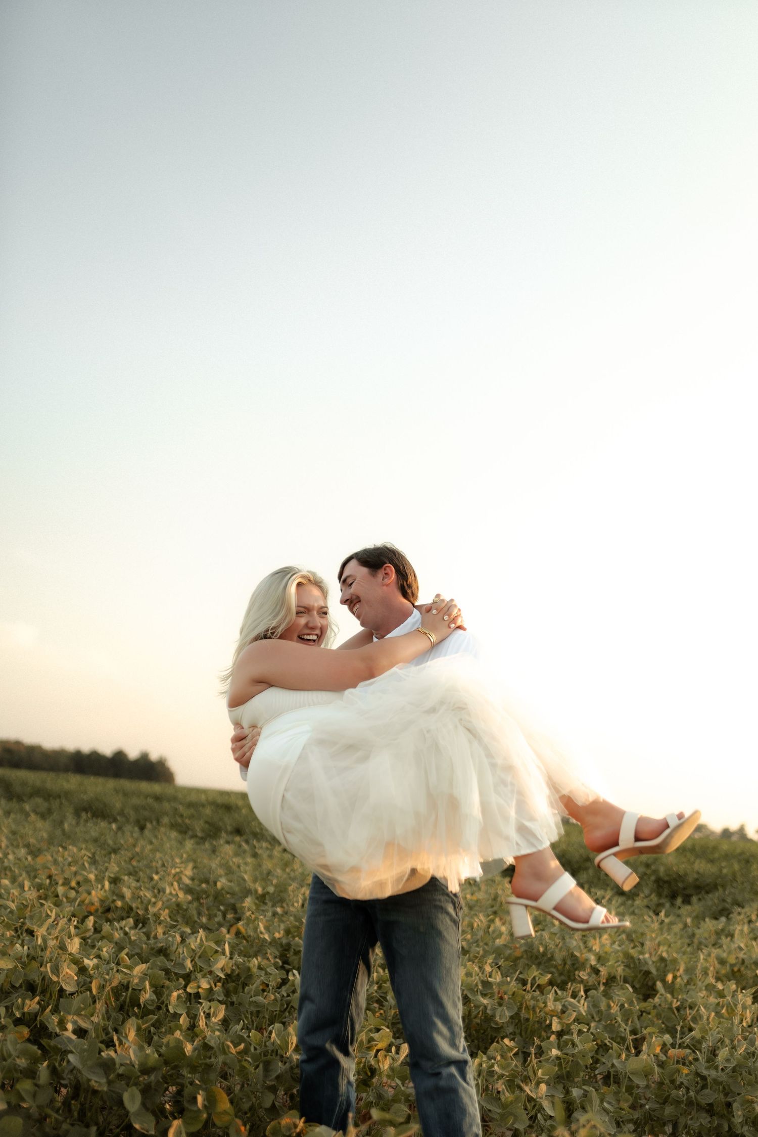 Groom lifting smiling bride in his arms in a green field under a clear sky.